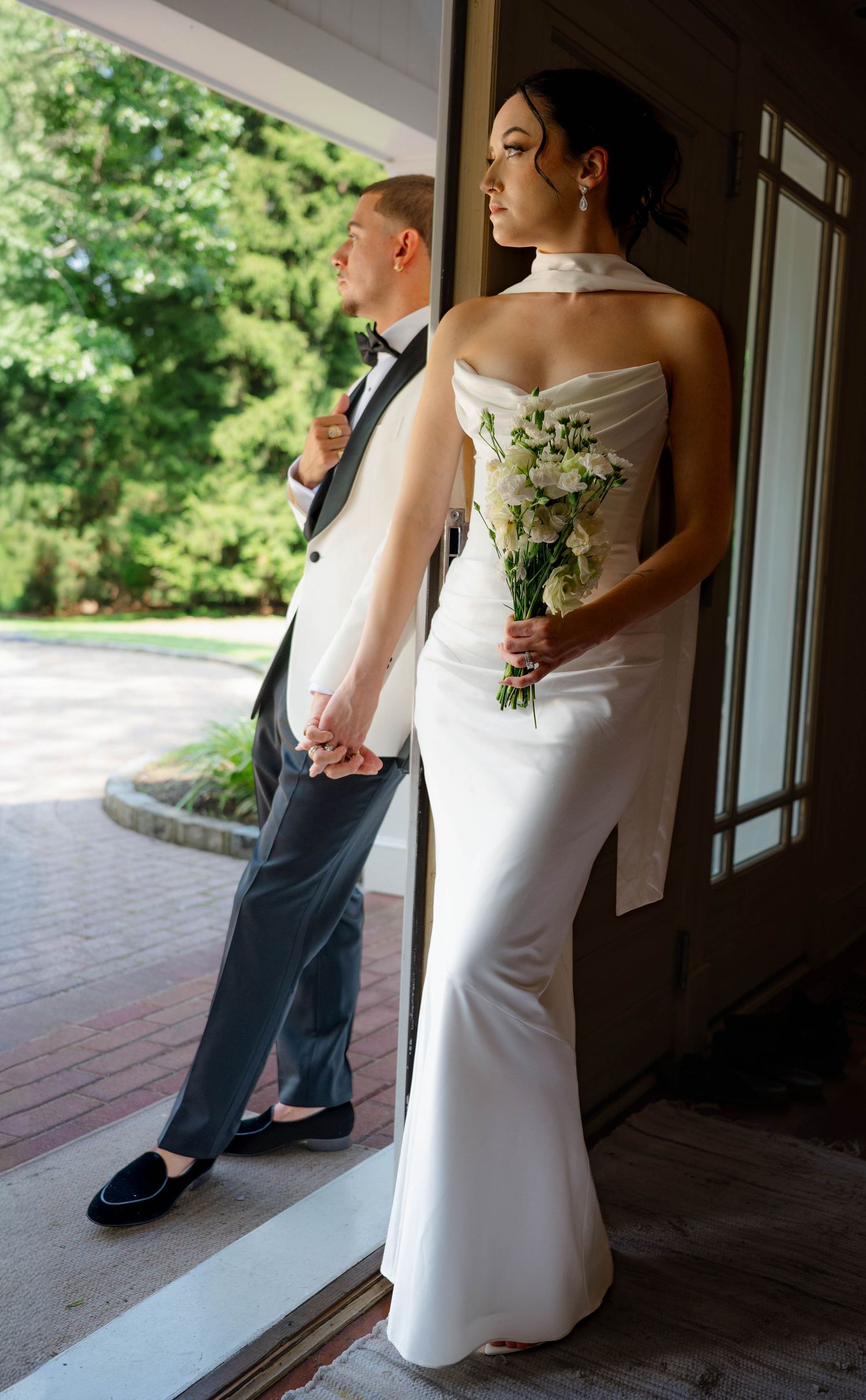 Bride in white halter gown holding bouquet stands with groom in gray tuxedo at venue entrance.