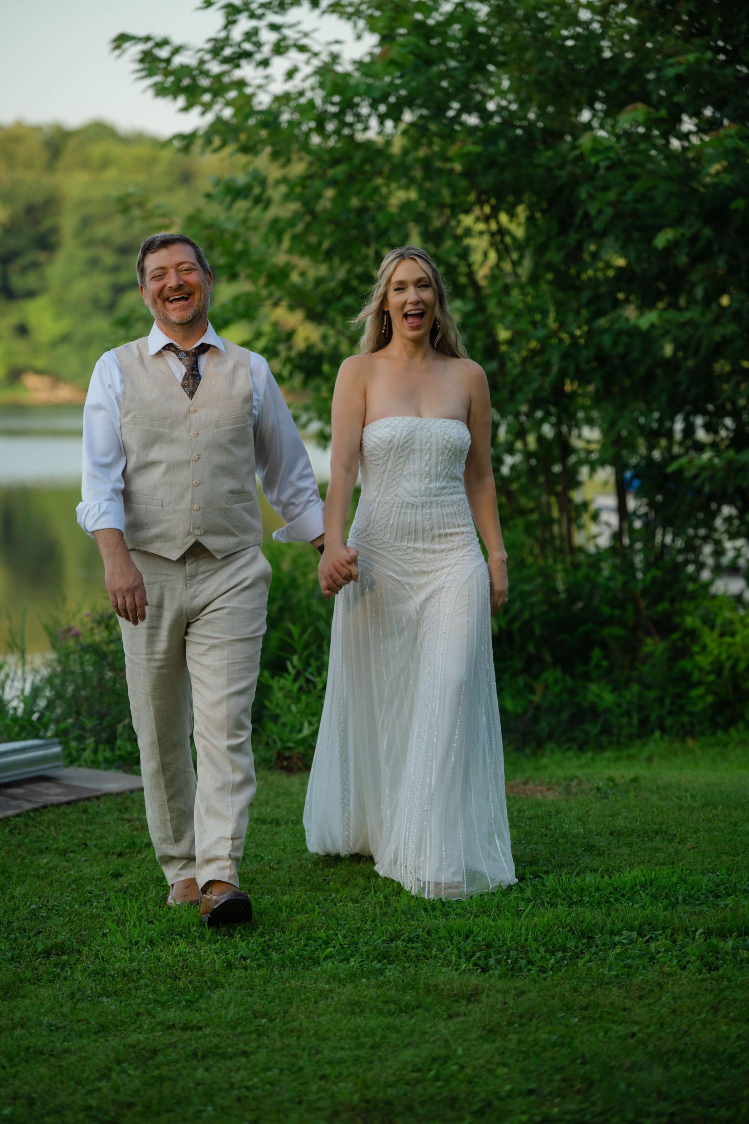 Happy bride and groom walking hand in hand on green grass near a lake, bride in strapless white gown, groom in tan vest.
