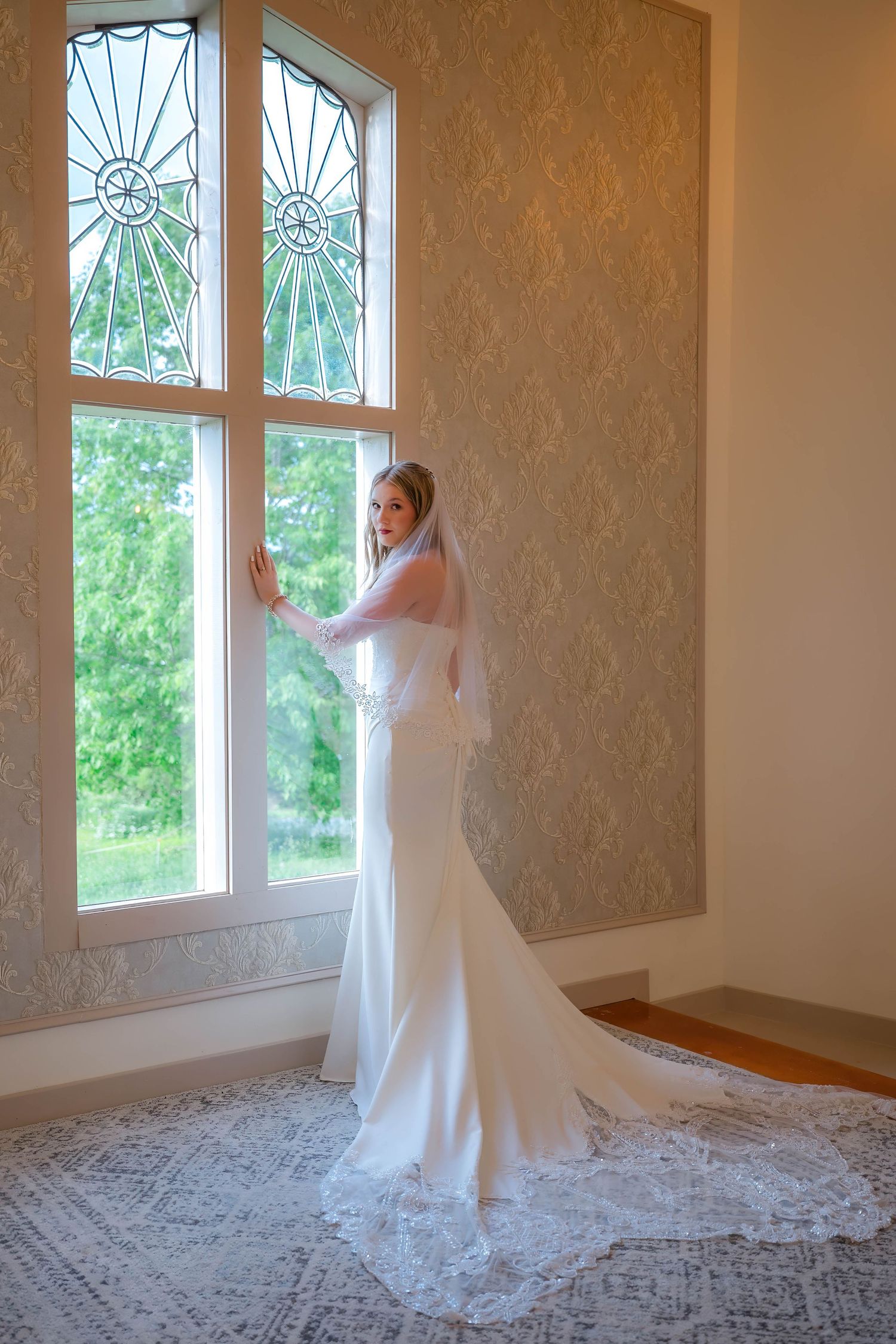 Elegant bride in white mermaid gown and lace veil stands by ornate stained glass window in softly lit room.