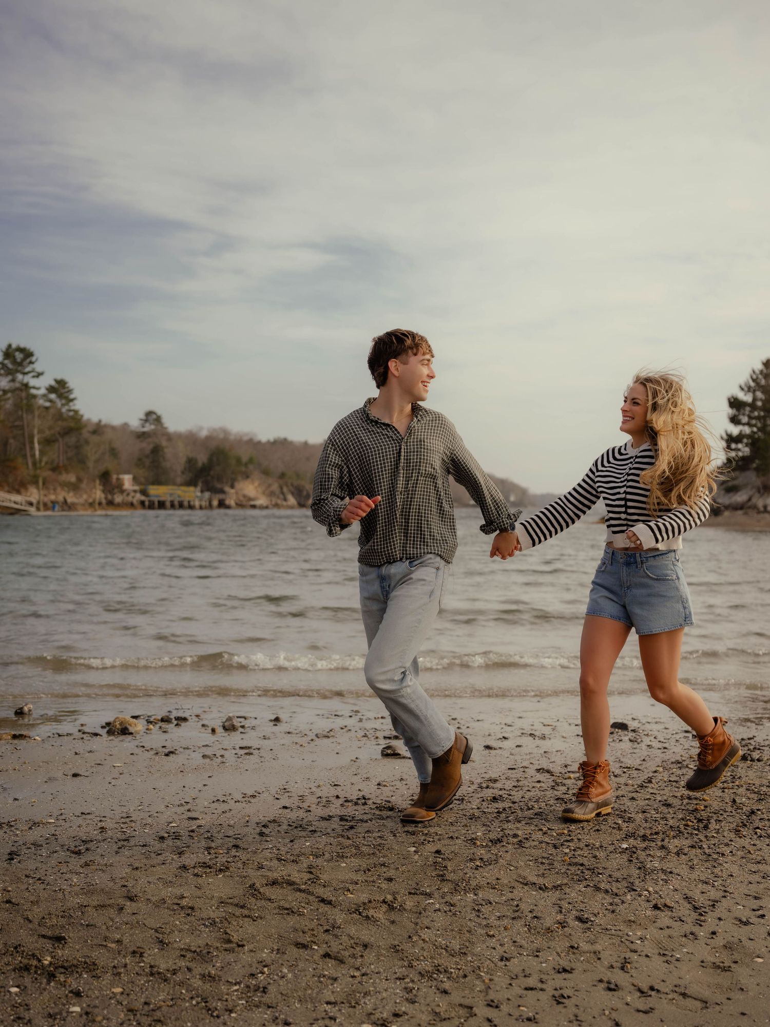 Happy couple running and holding hands along a scenic shoreline on a cloudy day.
