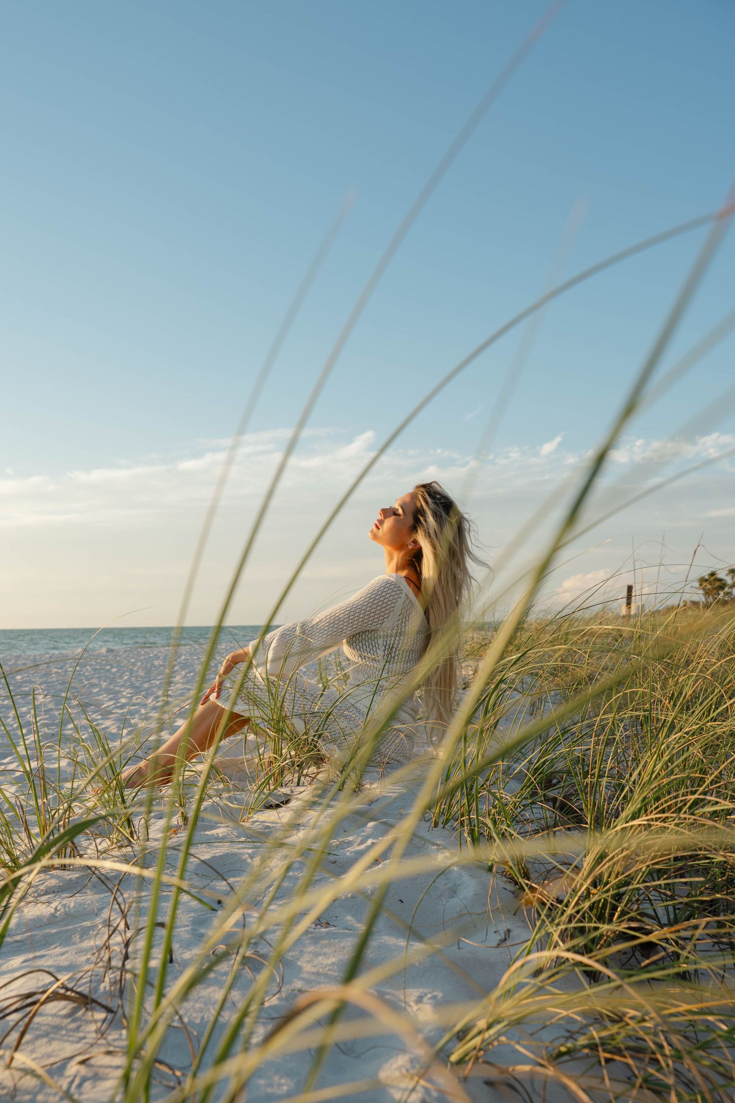 Blonde woman in a white dress sitting on a white sand dune with beach grass and the ocean in the background.