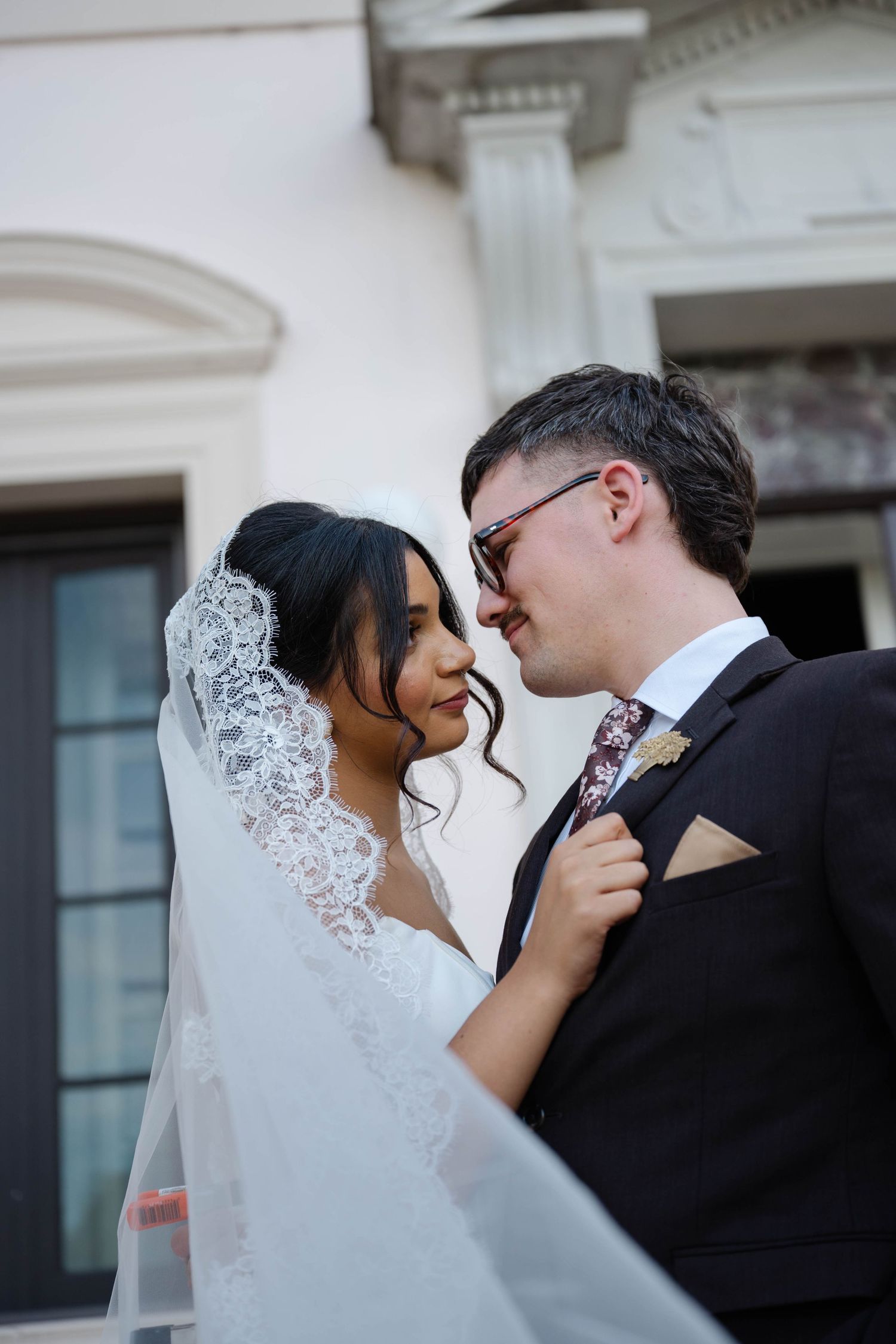 Bride and groom share a romantic close moment outside elegant white building, bride in lace veil.