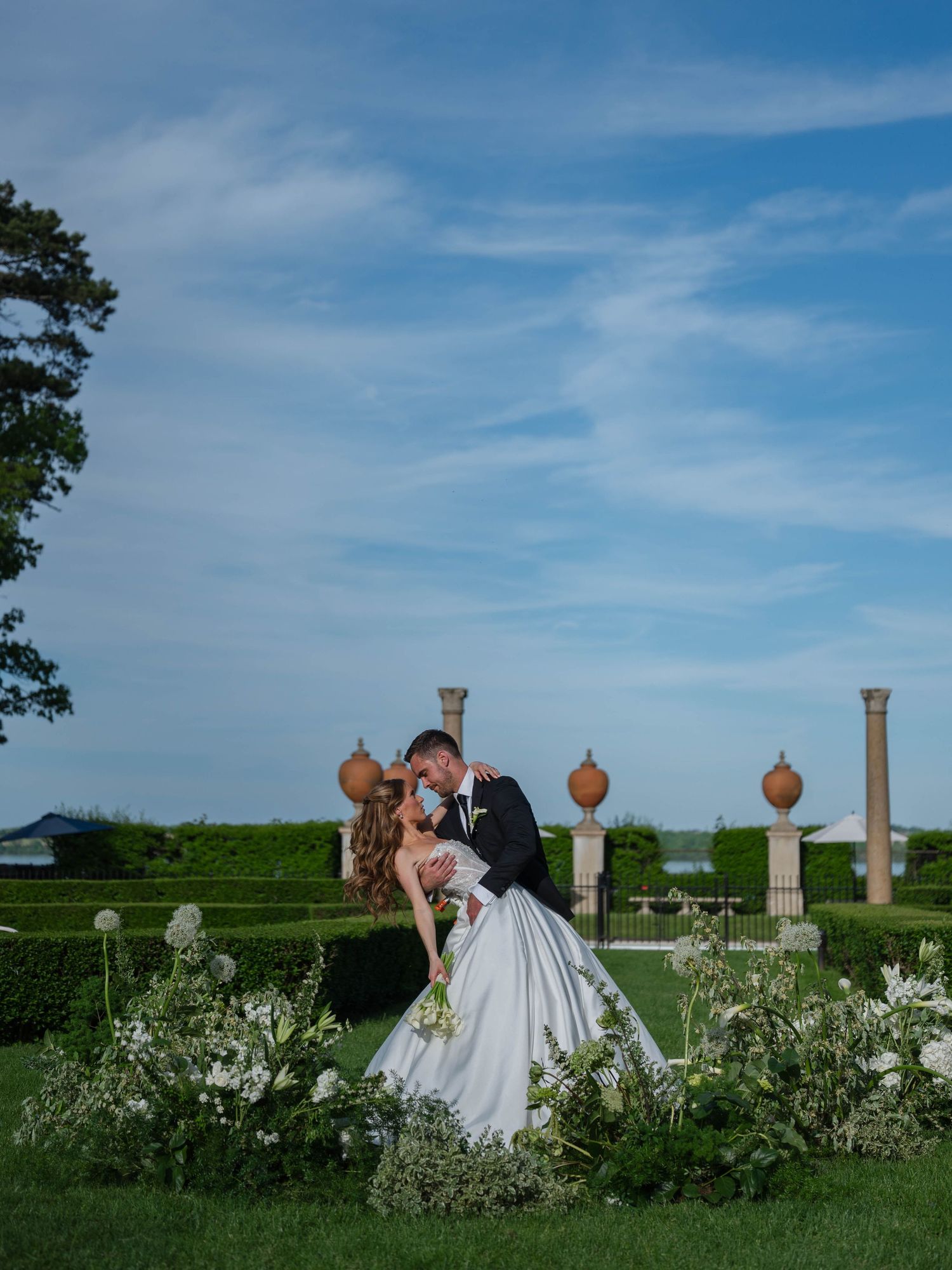 Bride and groom sharing romantic dip kiss in formal hedge garden with stone columns and blue sky.