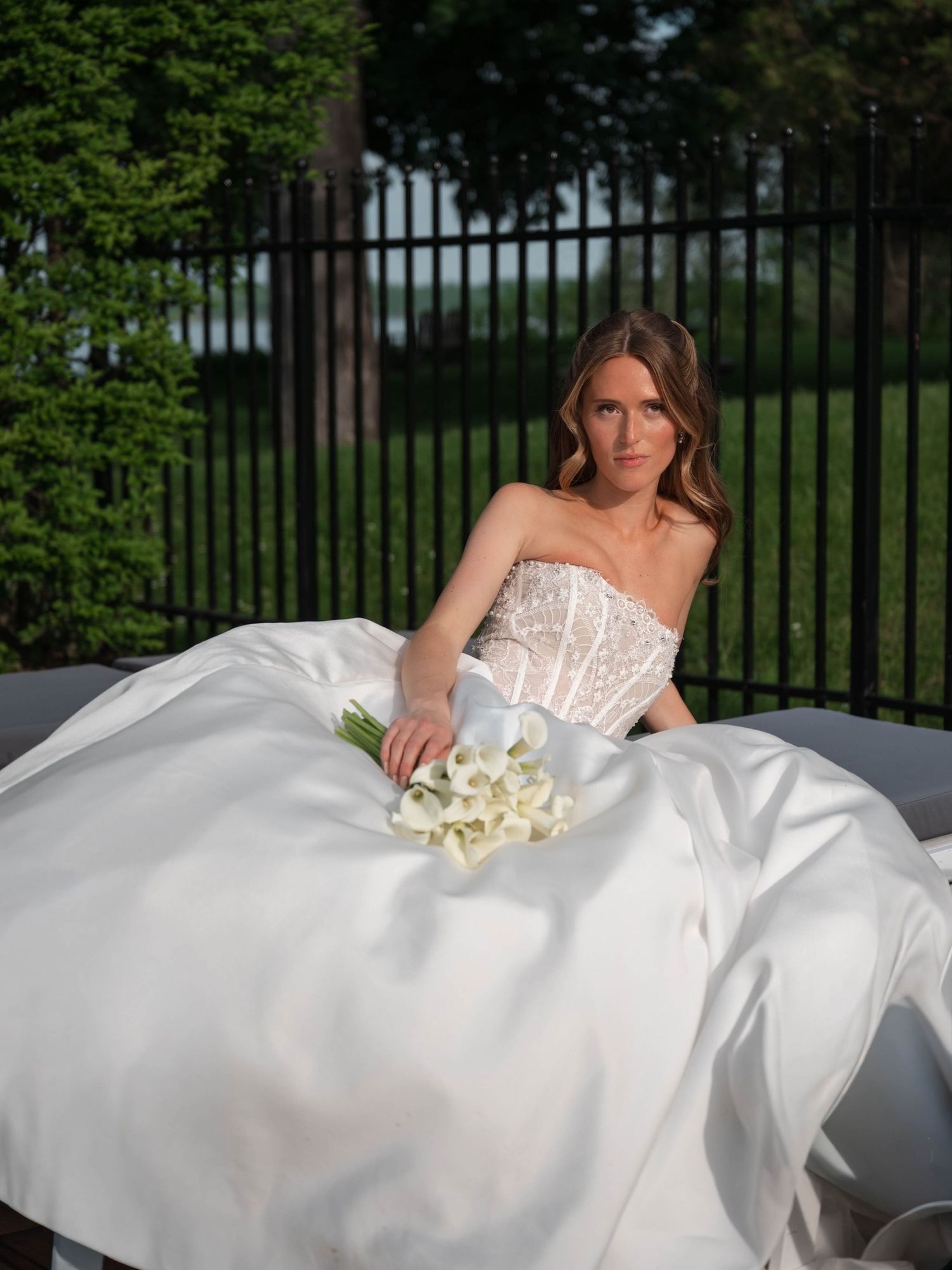 Elegant bride in beaded strapless ballgown holds calla lily bouquet while seated on outdoor lounge.