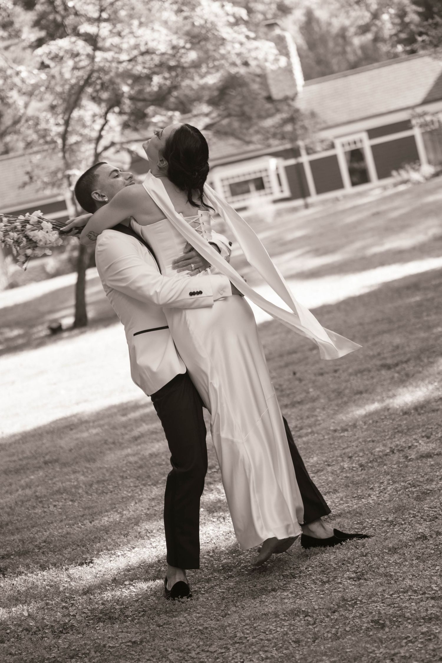 Black and white photo of a groom lifting his bride outdoors near trees and a building on their wedding day.