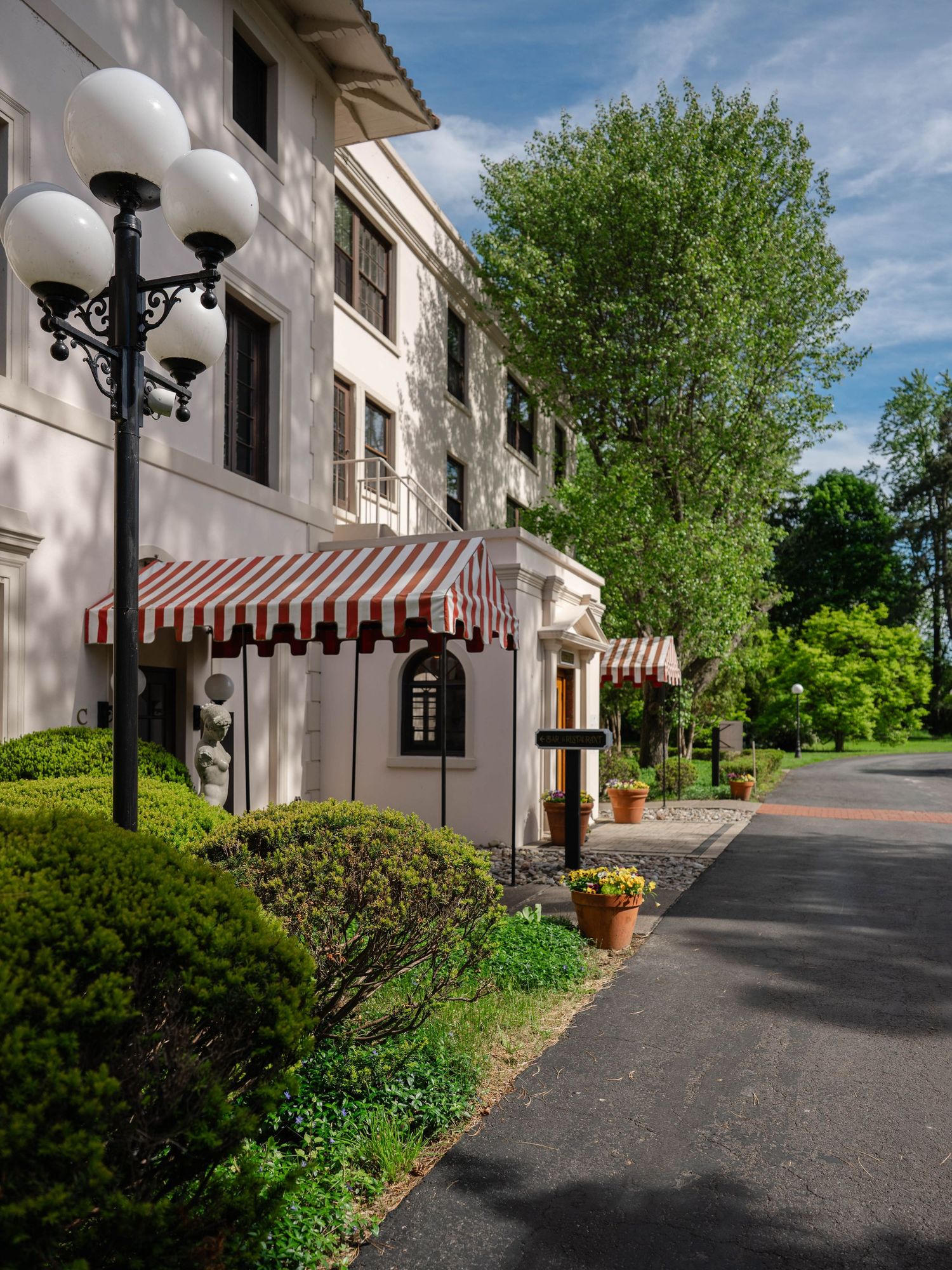 Elegant building exterior with red striped awnings, ornate lamp post, lush greenery, and a paved driveway on a sunny day.