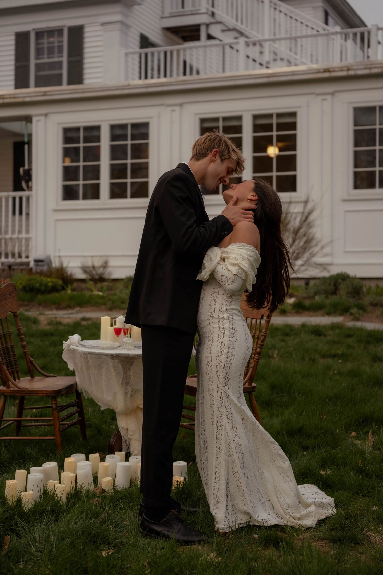 A couple shares a romantic kiss outdoors at dusk, surrounded by candles, the bride in a lace gown and groom in a black suit.