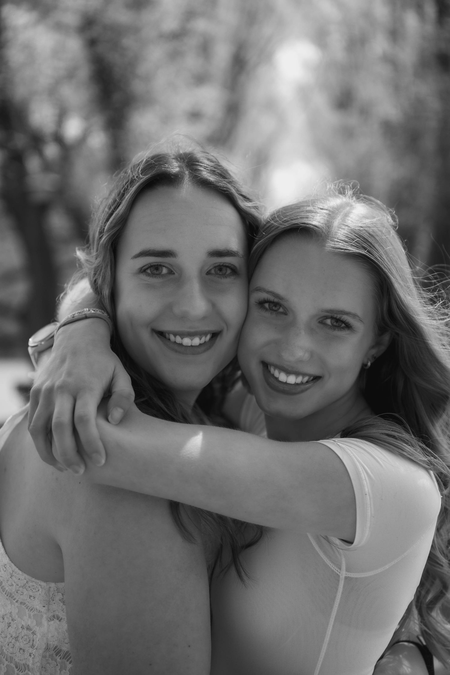 Black and white portrait of two smiling young women embracing warmly in an outdoor park setting.