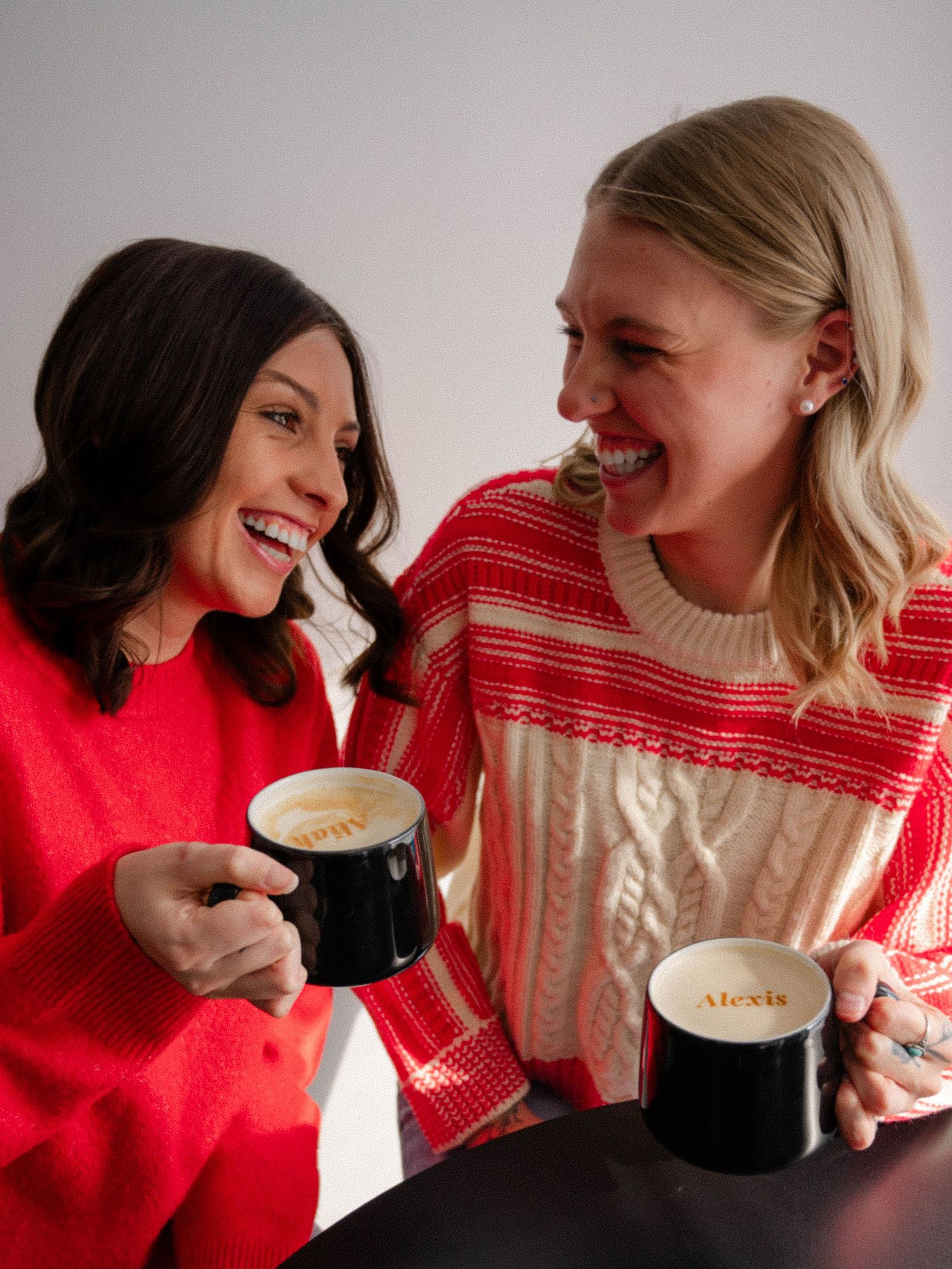 Two women laughing together holding personalized mugs in festive holiday sweaters indoors.