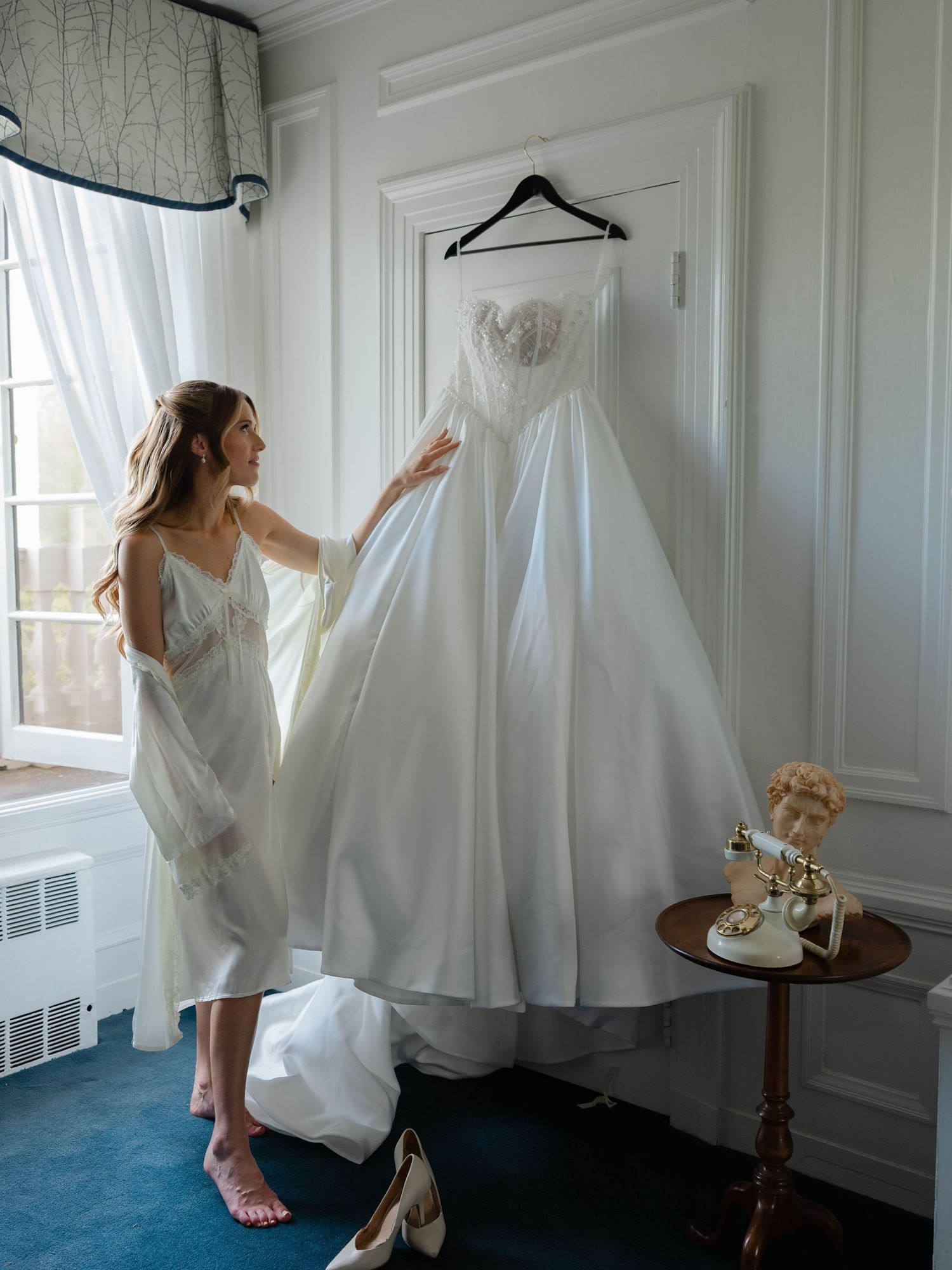 Bride in silk robe admires strapless ballgown wedding dress hanging in elegant hotel bridal suite.