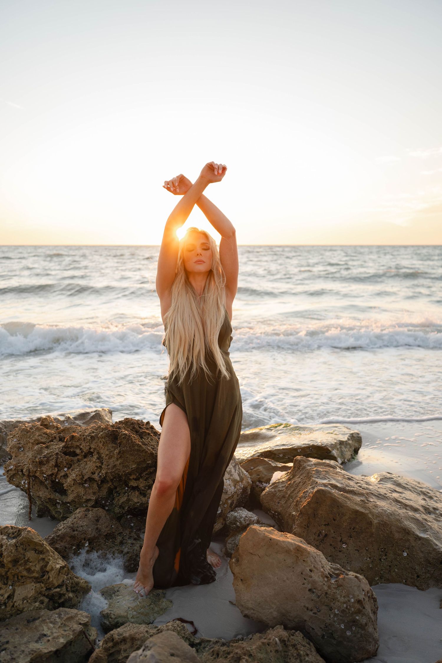 Blonde woman in olive dress poses with arms raised on rocky beach at golden sunset.