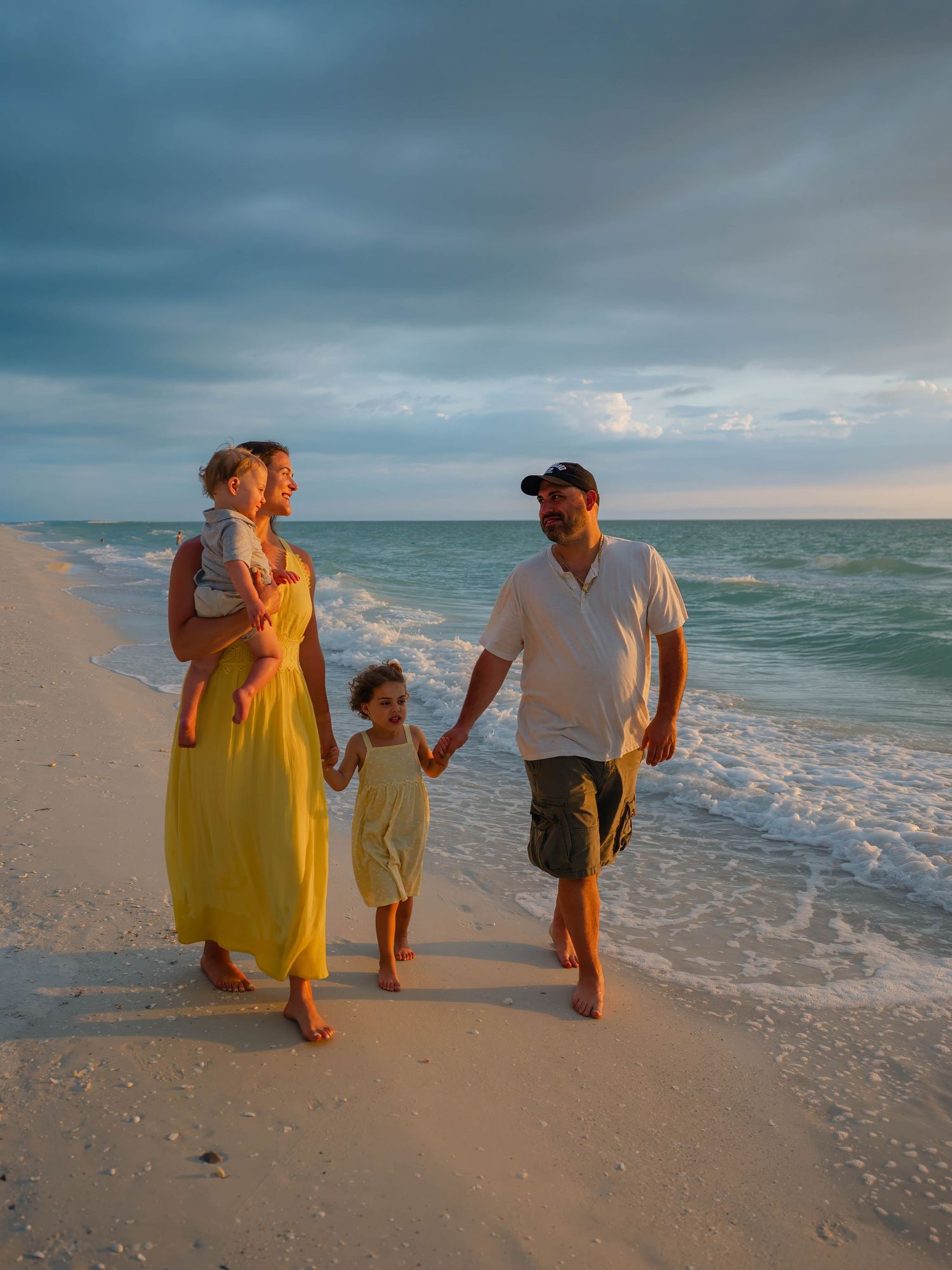 Happy family walking on white sand beach at golden hour, mother in yellow dress holding baby.