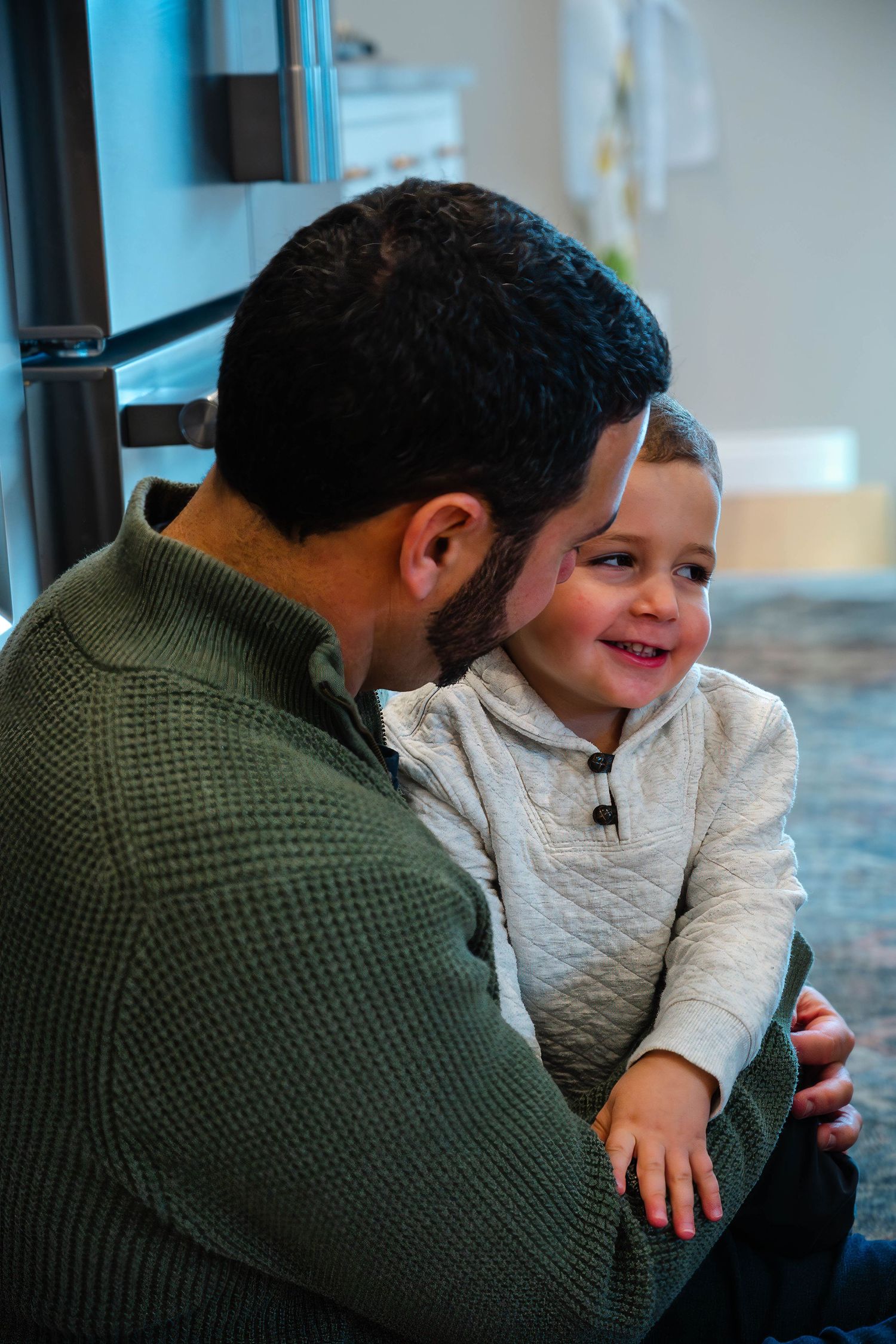 A bearded father in a green sweater holds his smiling toddler son in a bright kitchen.