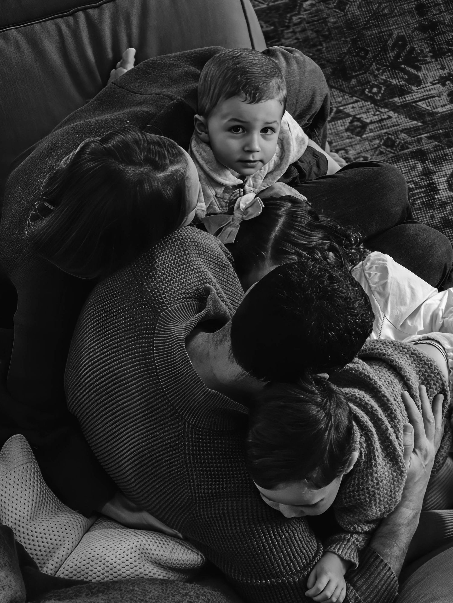 Family cuddles together on a couch in a candid black and white photo with young child looking at camera.