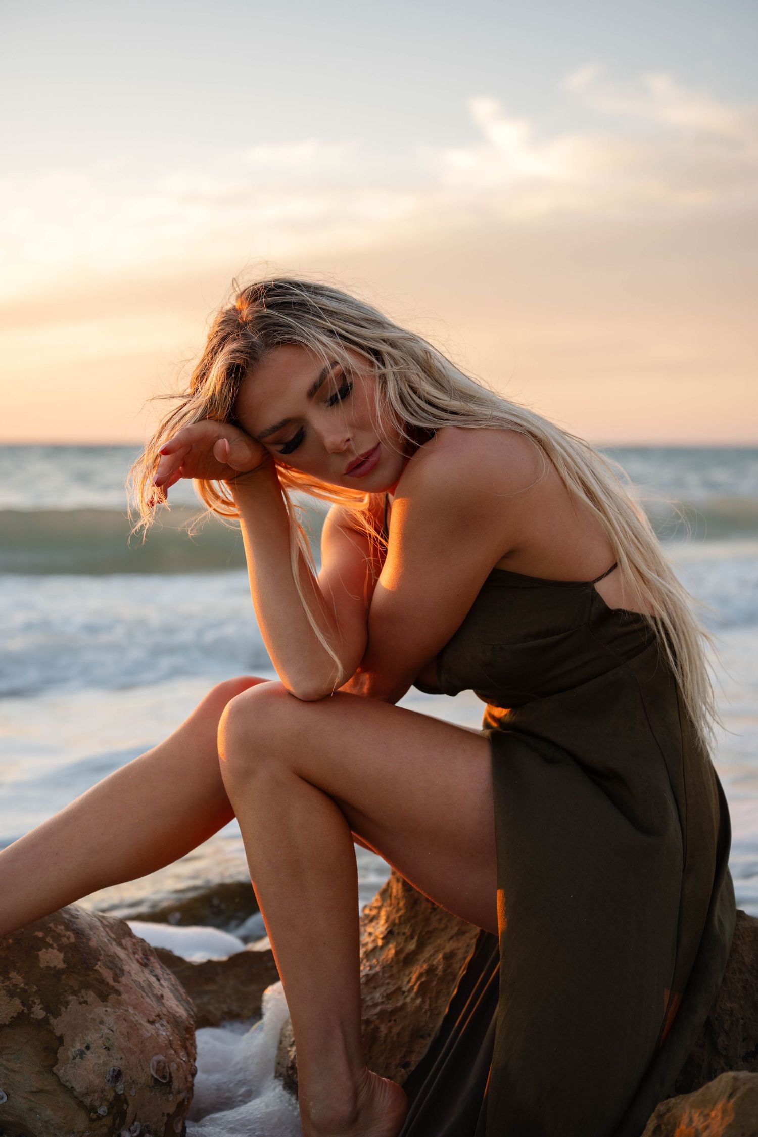 Blonde woman in olive dress sits on beach rocks in contemplative pose at sunset.