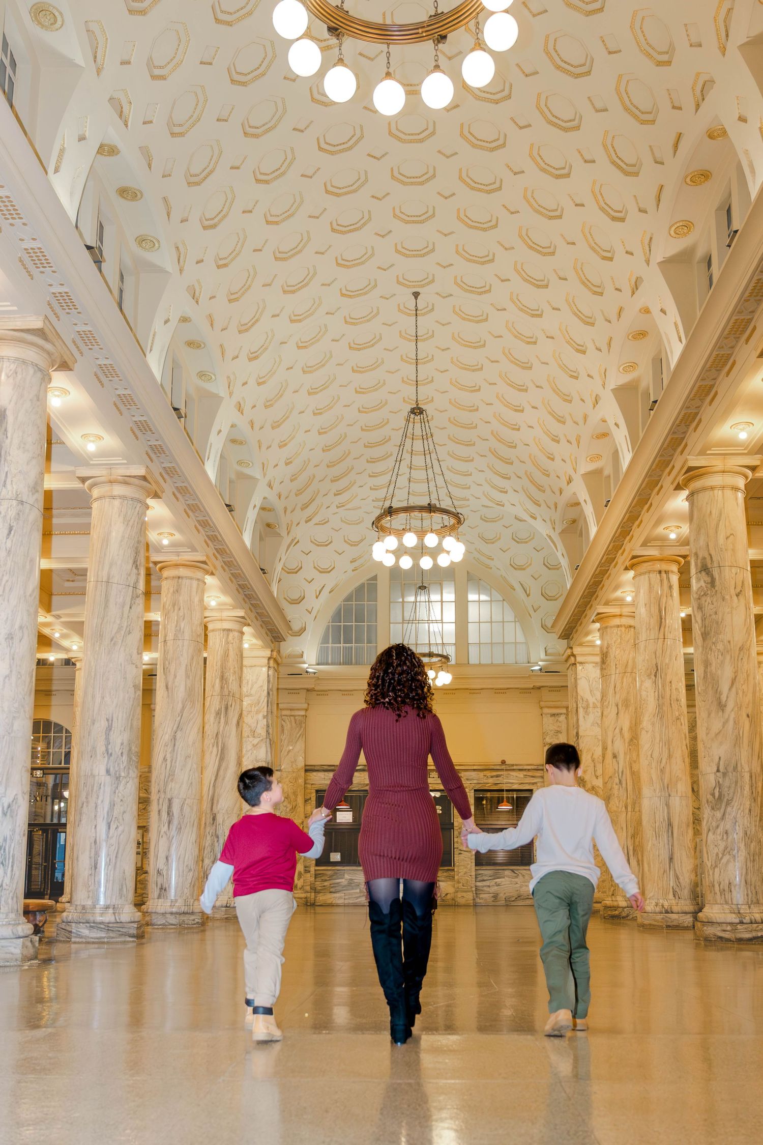 Pregnant mother walks hand in hand with two sons through grand marble hall with ornate ceiling.