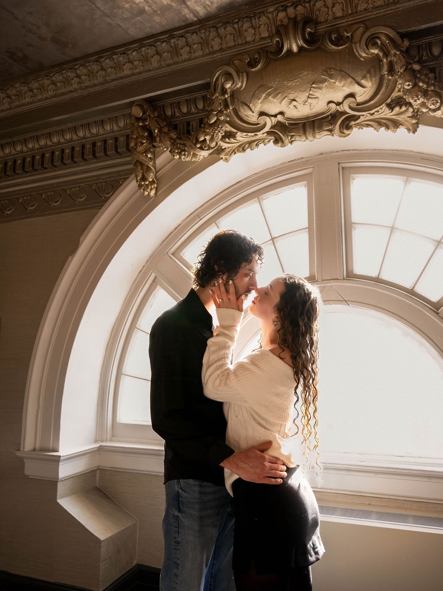 Couple sharing a romantic kiss in front of an ornate arched window in an elegant historic building.