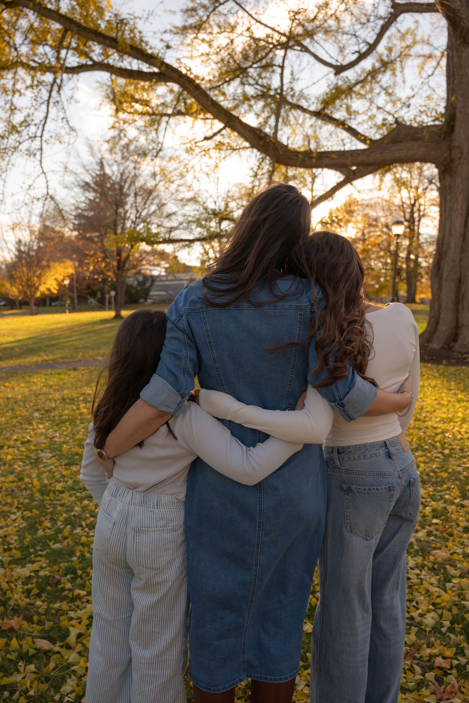 Mother and two daughters hugging from behind under large tree in scenic autumn park at sunset.
