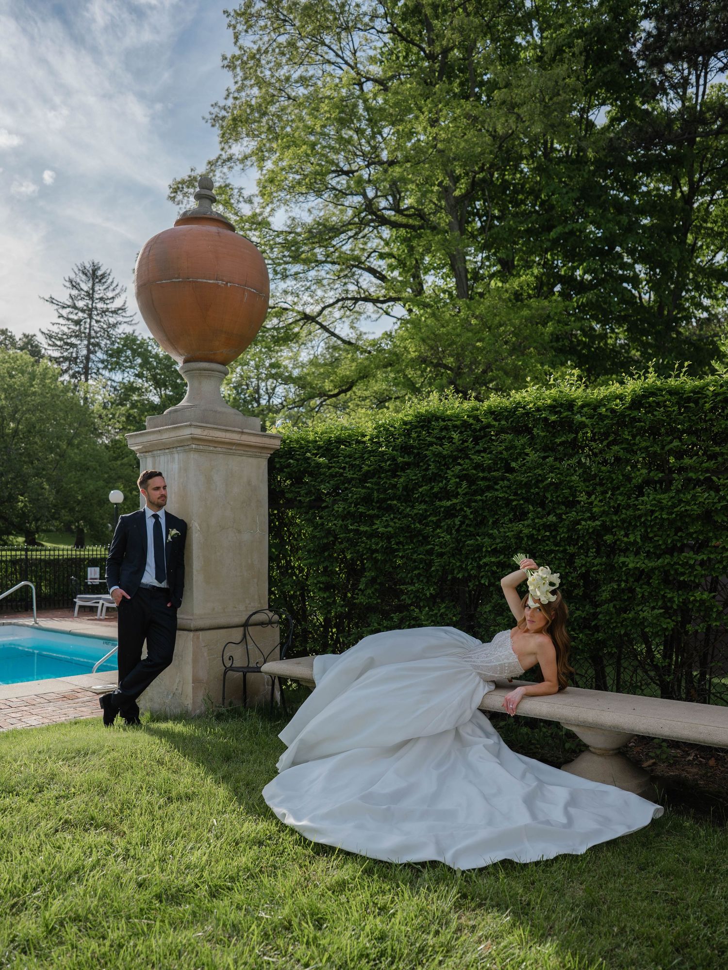 Bride in full ball gown reclining on stone bench near pool as groom stands by ornate garden pillar.