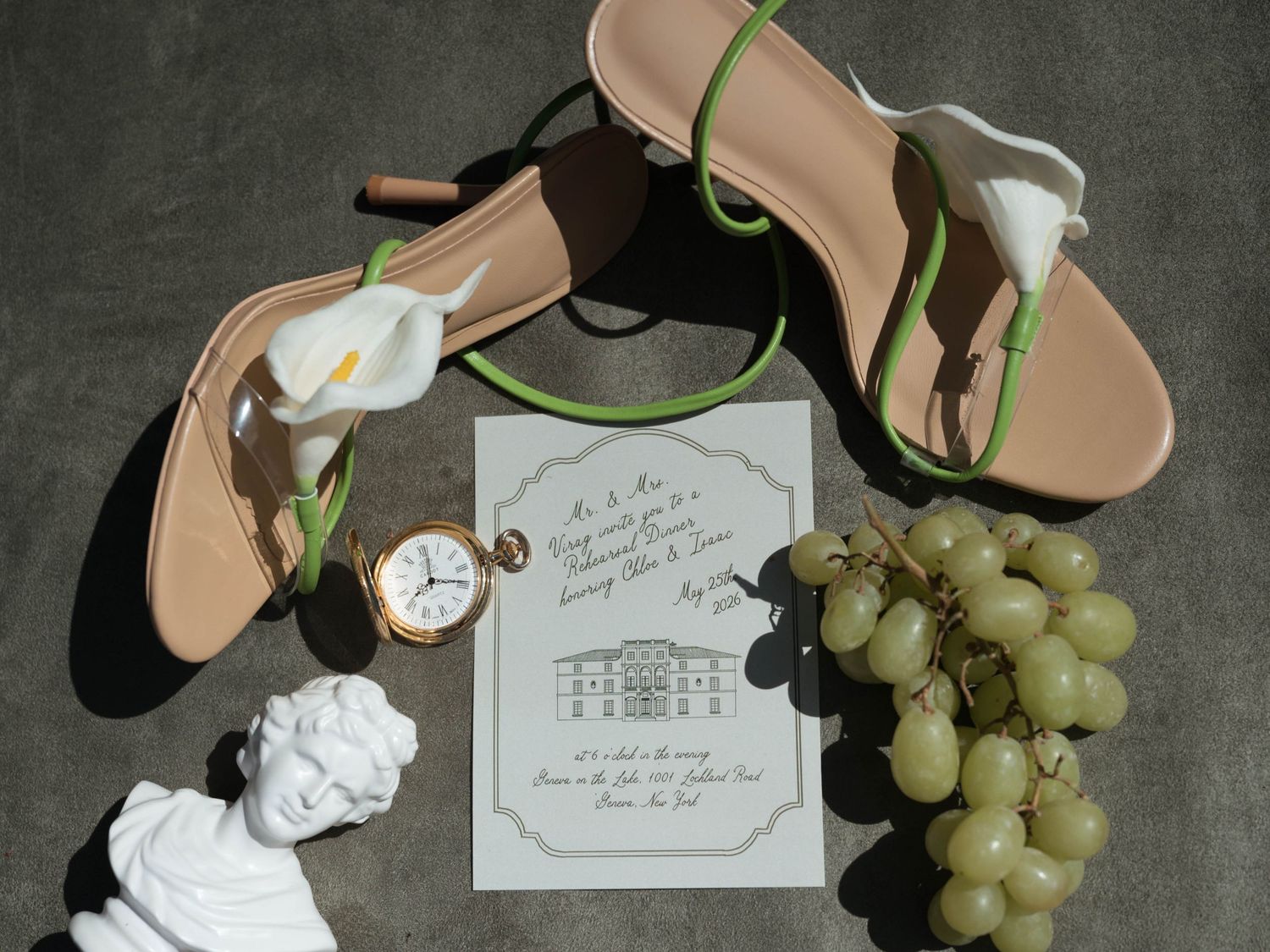 Flat lay of wedding invitation, sandals, pocket watch, grapes, and white calla lily on gray surface.