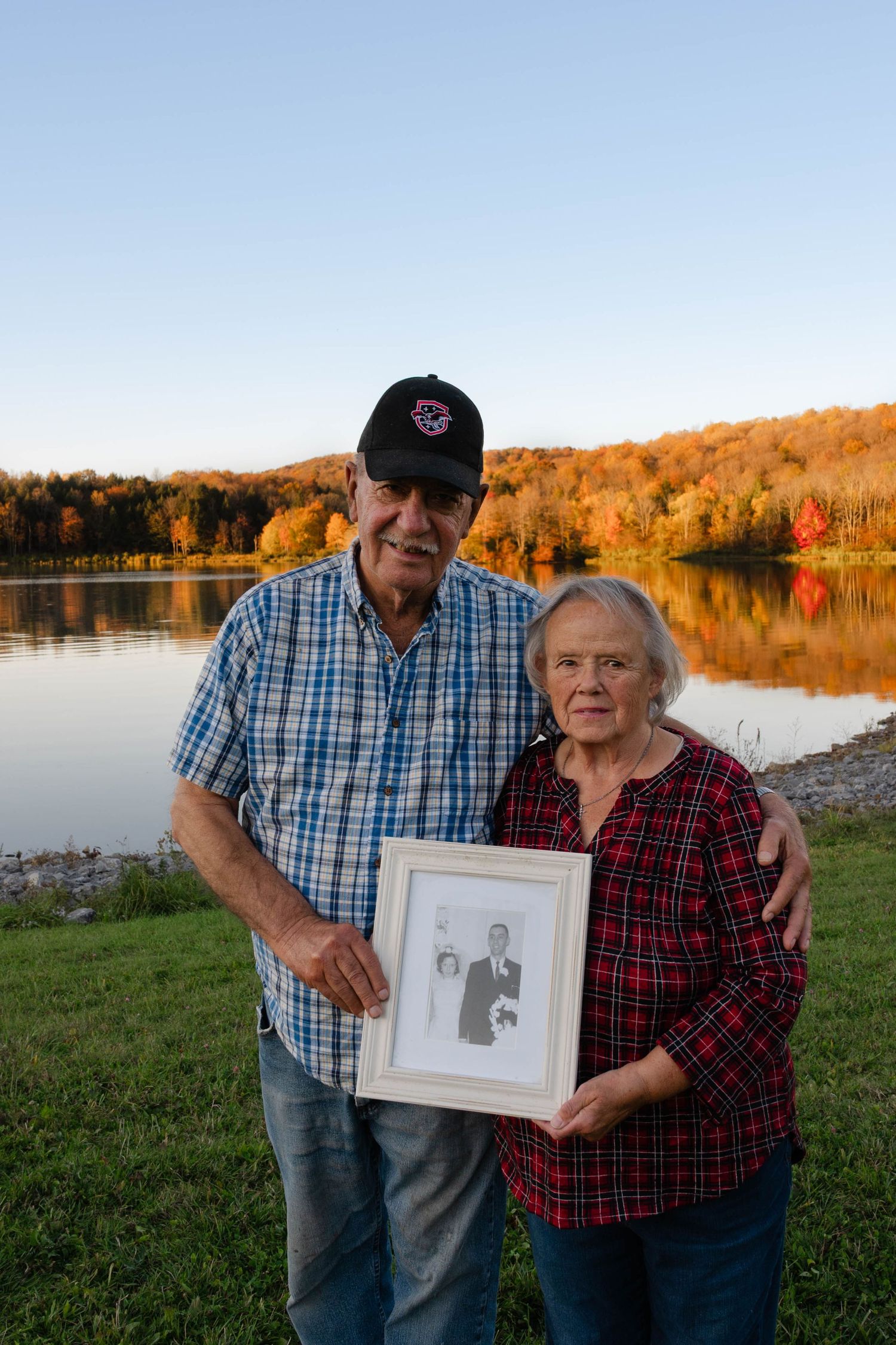 Senior couple holding a framed wedding photo by a lake with stunning autumn foliage in background.