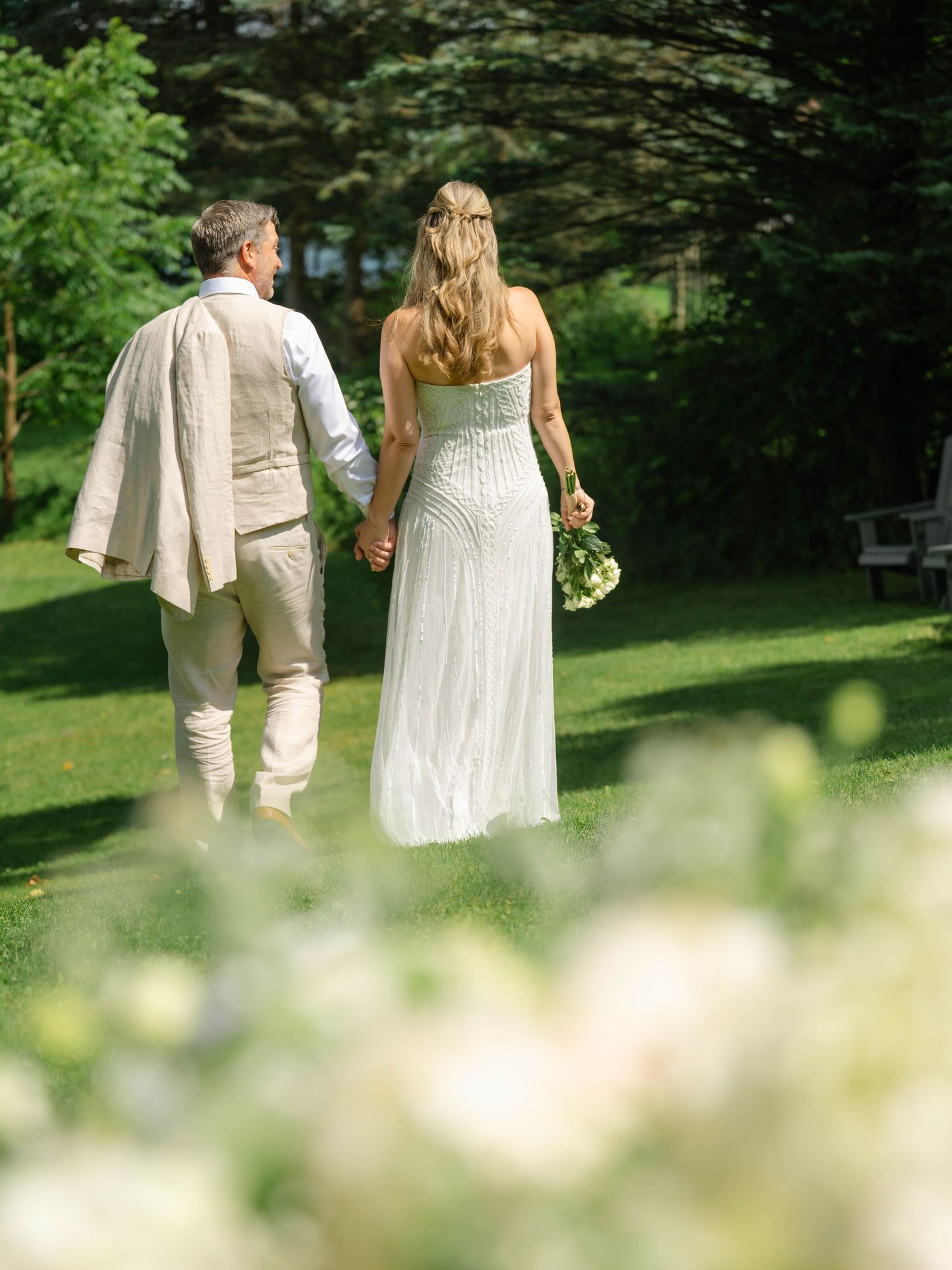 A bride and groom walk hand-in-hand through a lush green garden, she in a white gown holding a bouquet.