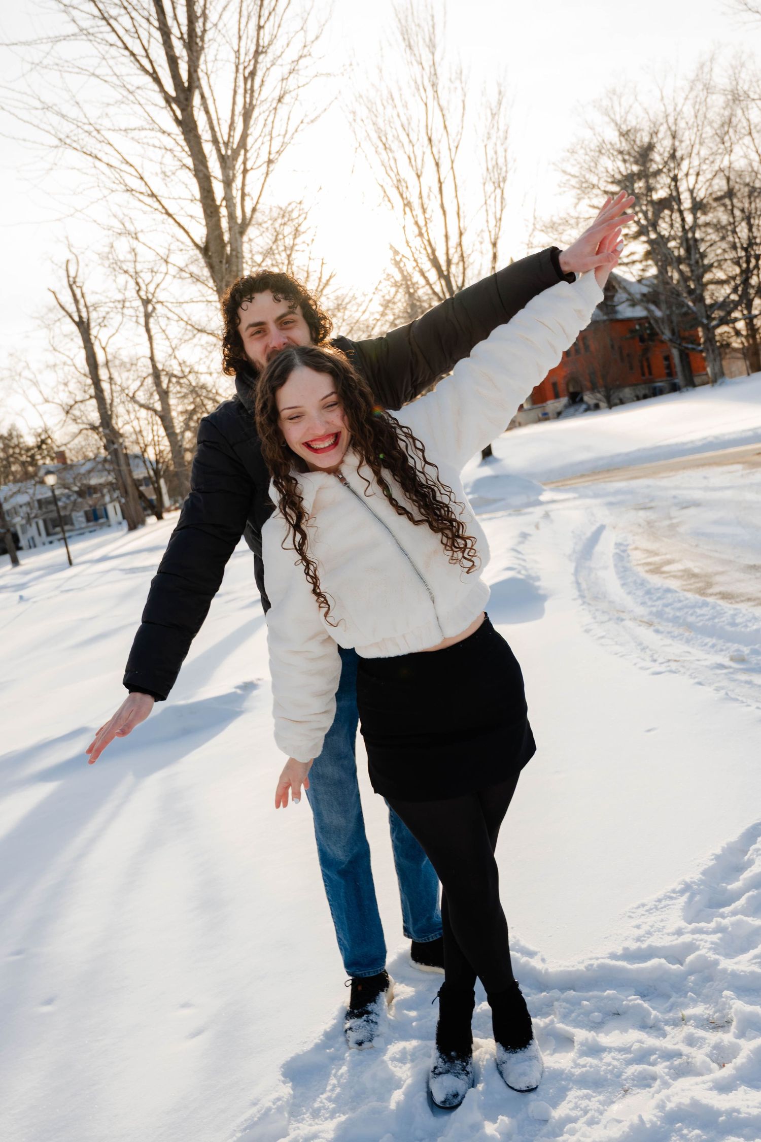 Happy couple playing in snow, woman laughing with arms outstretched in sunny winter park.