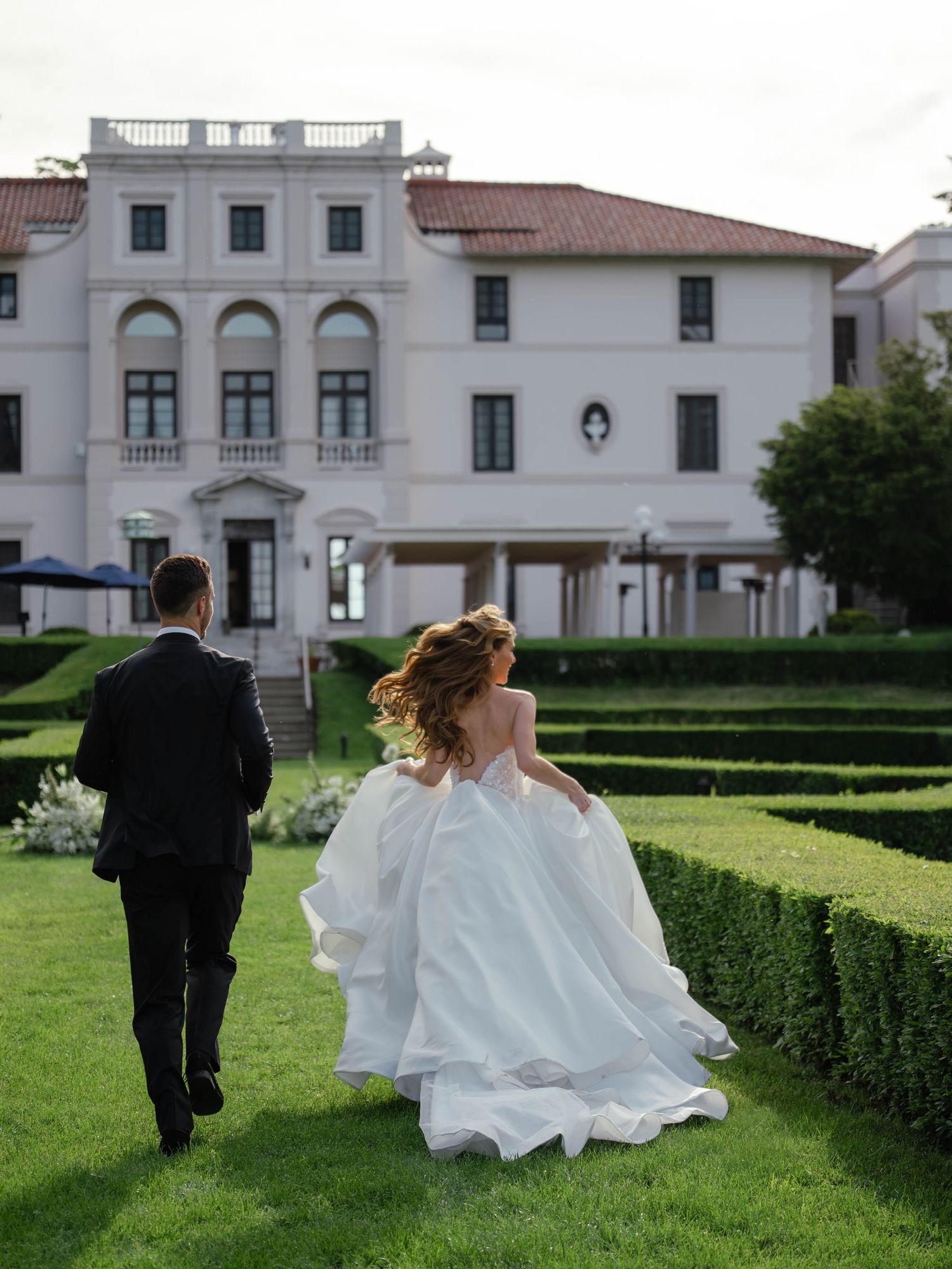 Bride in flowing white ball gown and groom walking toward elegant white mansion with hedge maze.