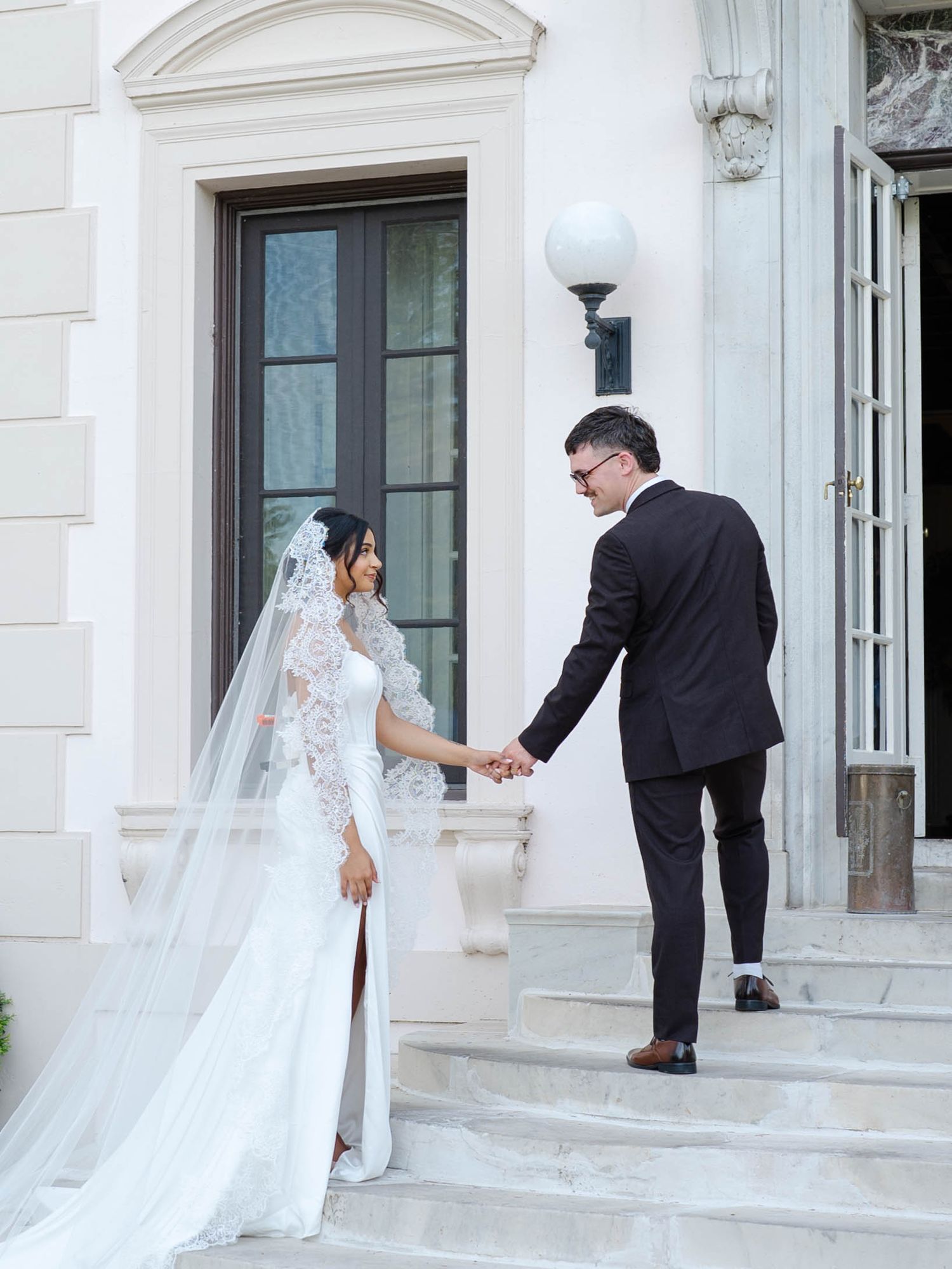 Wedding couple holds hands on steps of grand white building, bride in lace veil gown facing groom. Geneva On The Lake