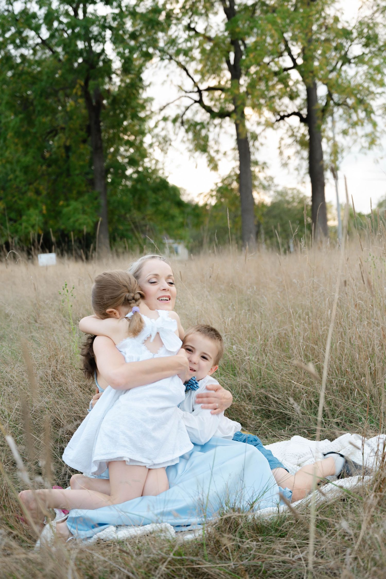 A smiling mother in white dress embraces her two young children while sitting in tall grass outdoors.