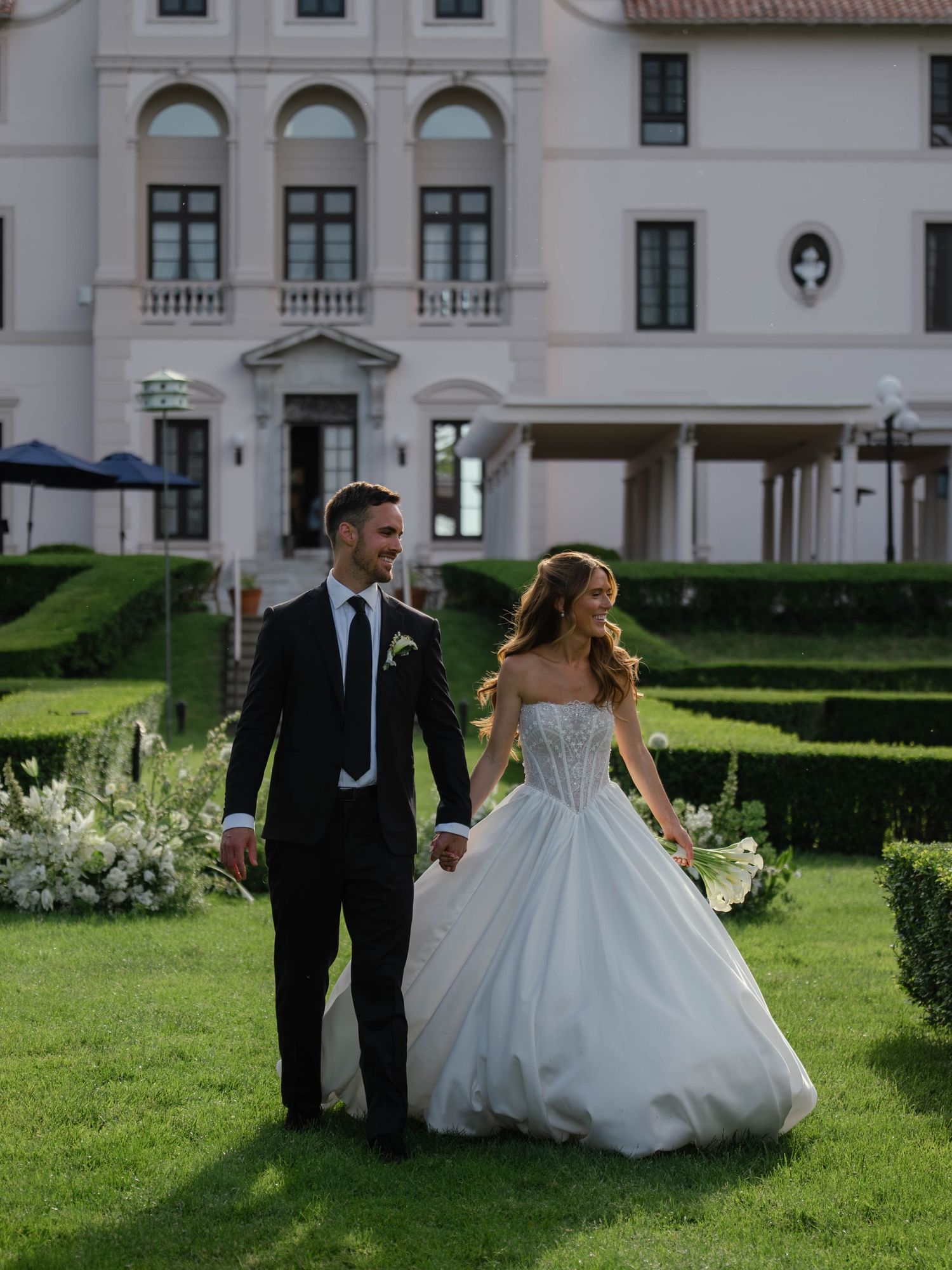 Smiling newlyweds holding hands walking on green lawn in front of elegant white mansion venue.