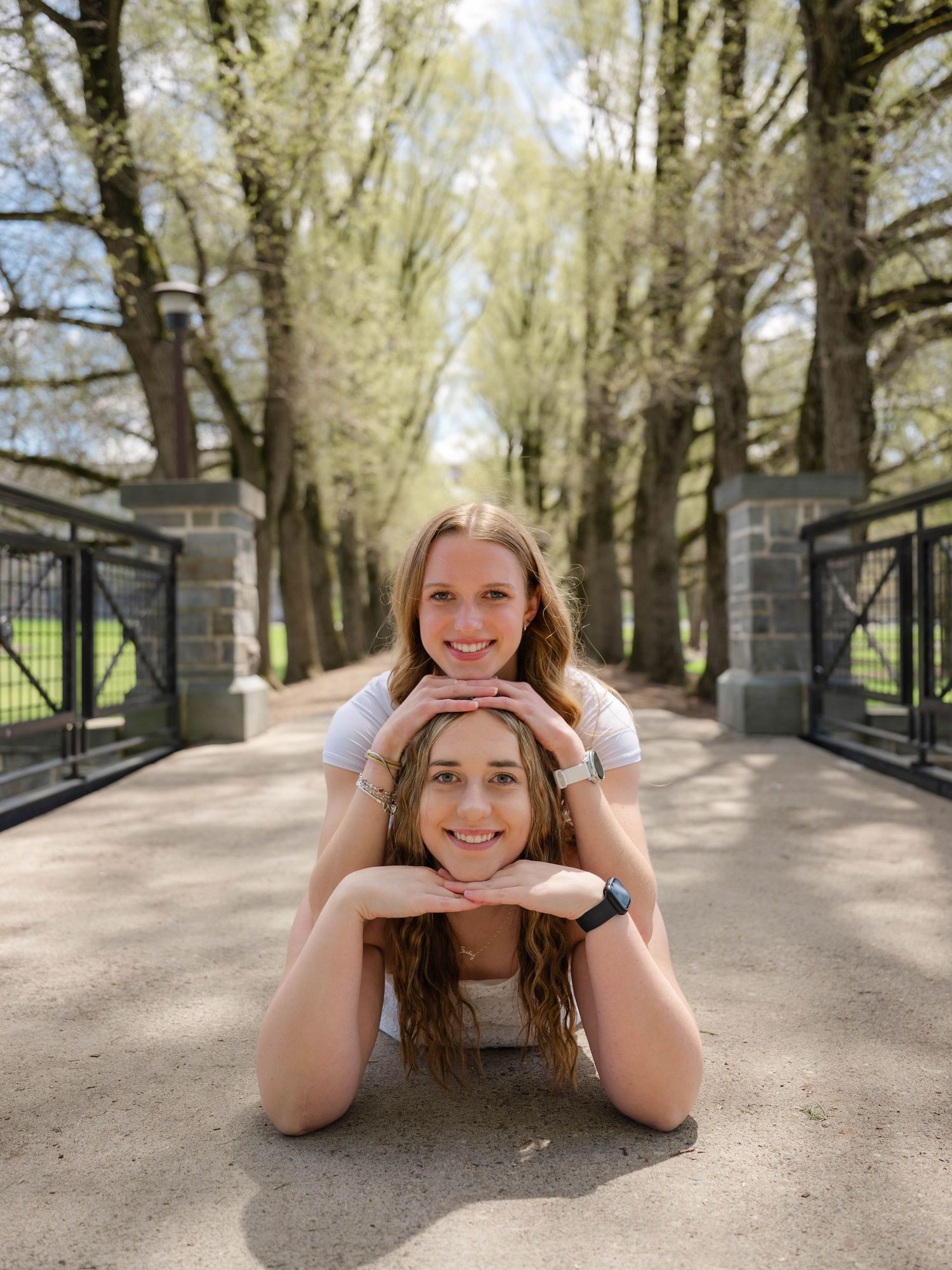 Two smiling young women pose playfully on a sunny tree-lined path, one lying down and one resting on her back.