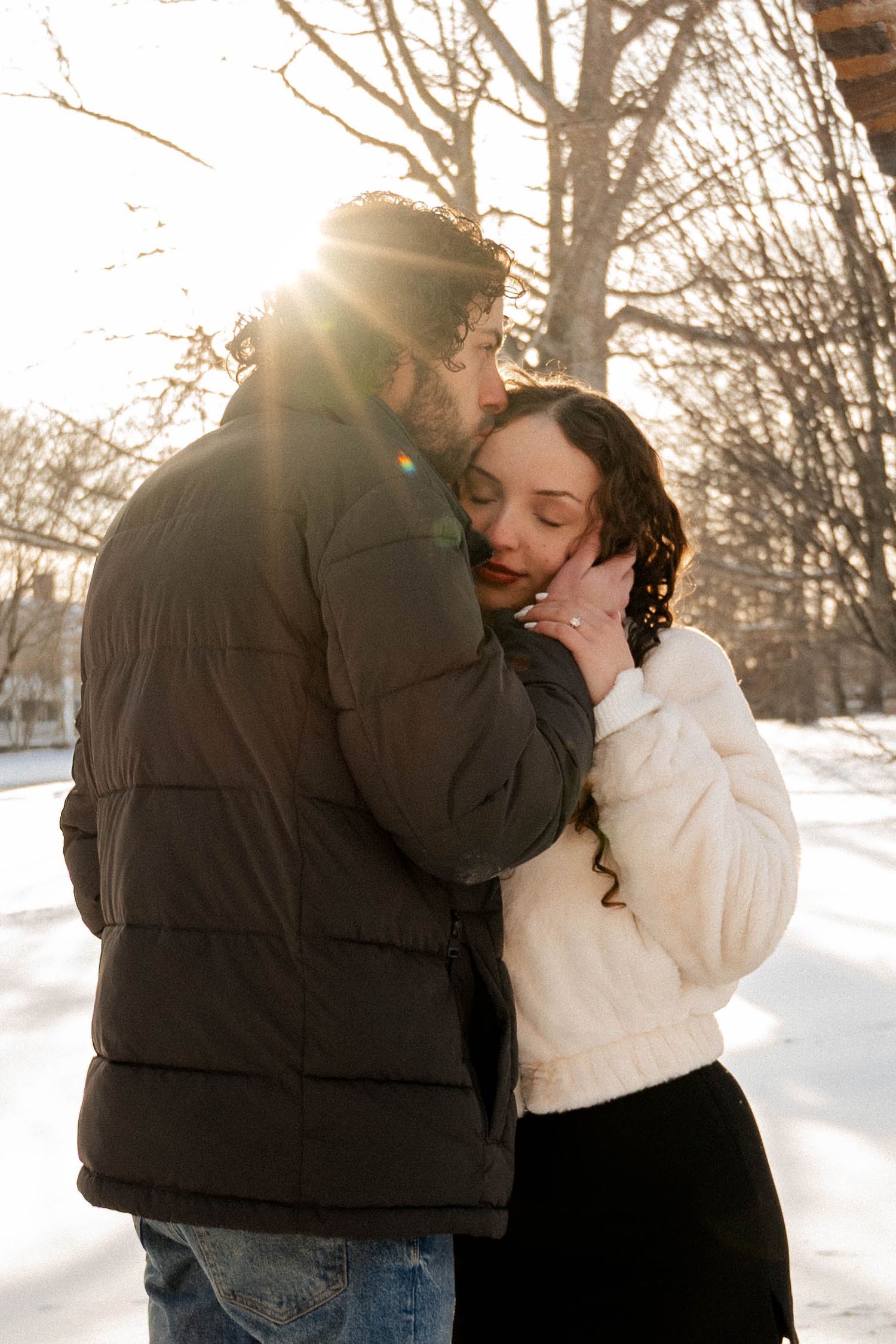 Man in dark puffer jacket embracing woman in white fur coat in sunny snowy park.