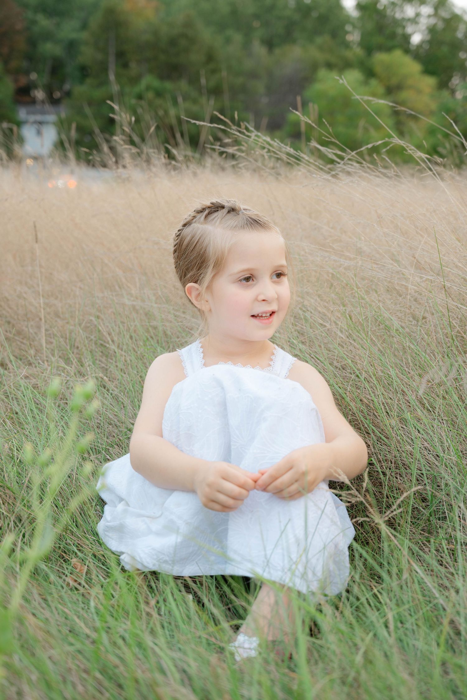 Adorable young girl in white dress sitting happily among tall golden grass in a natural outdoor setting.