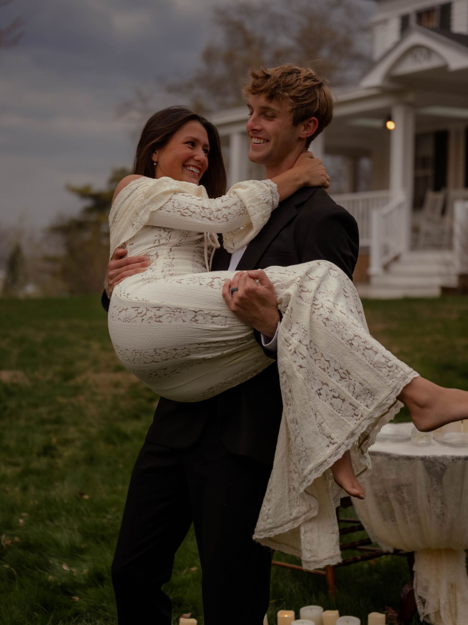 Joyful groom in black suit lifts smiling bride in lace wedding dress outdoors at dusk.