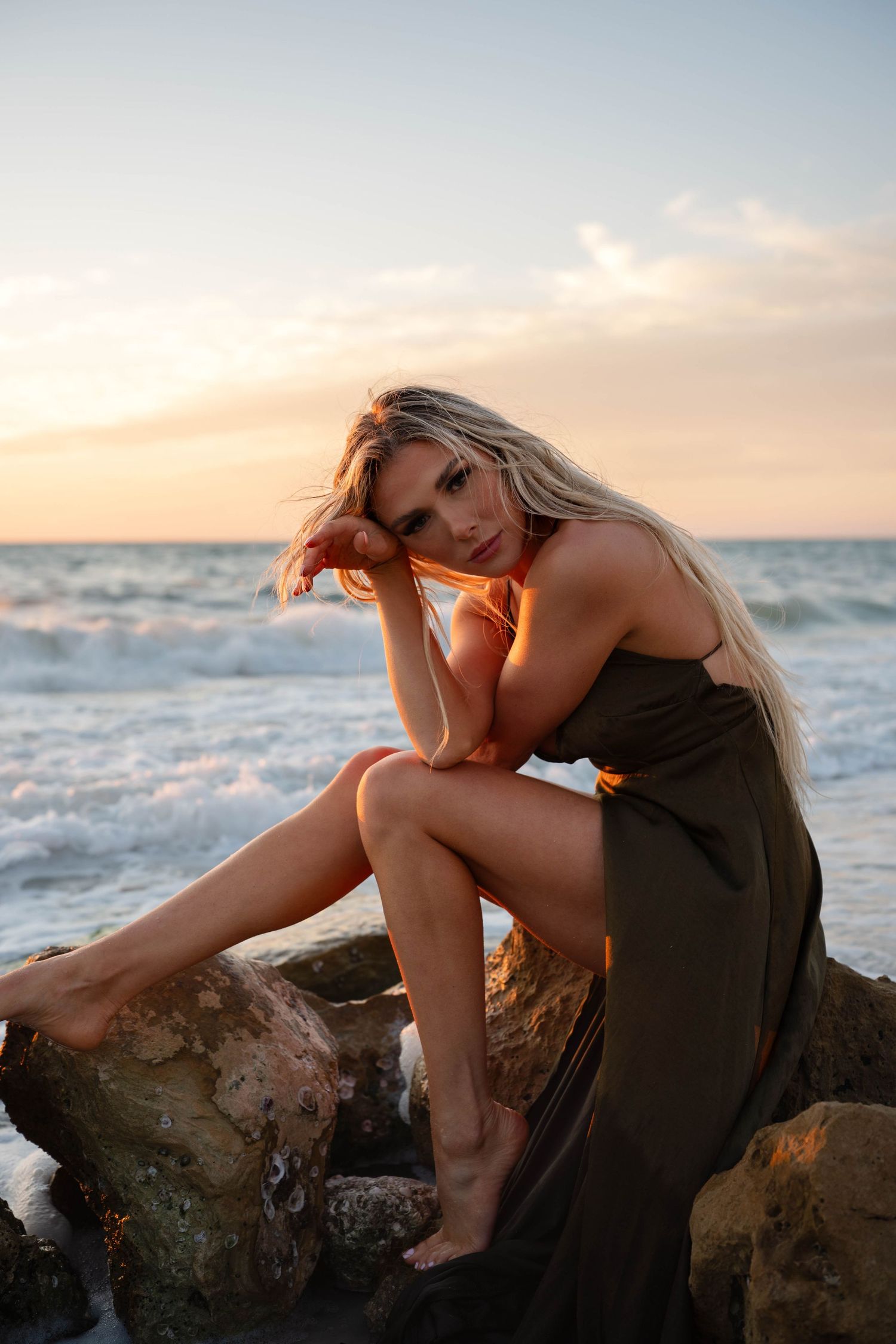 Blonde woman in olive dress sitting on rocks at sunset beach with ocean waves in background.