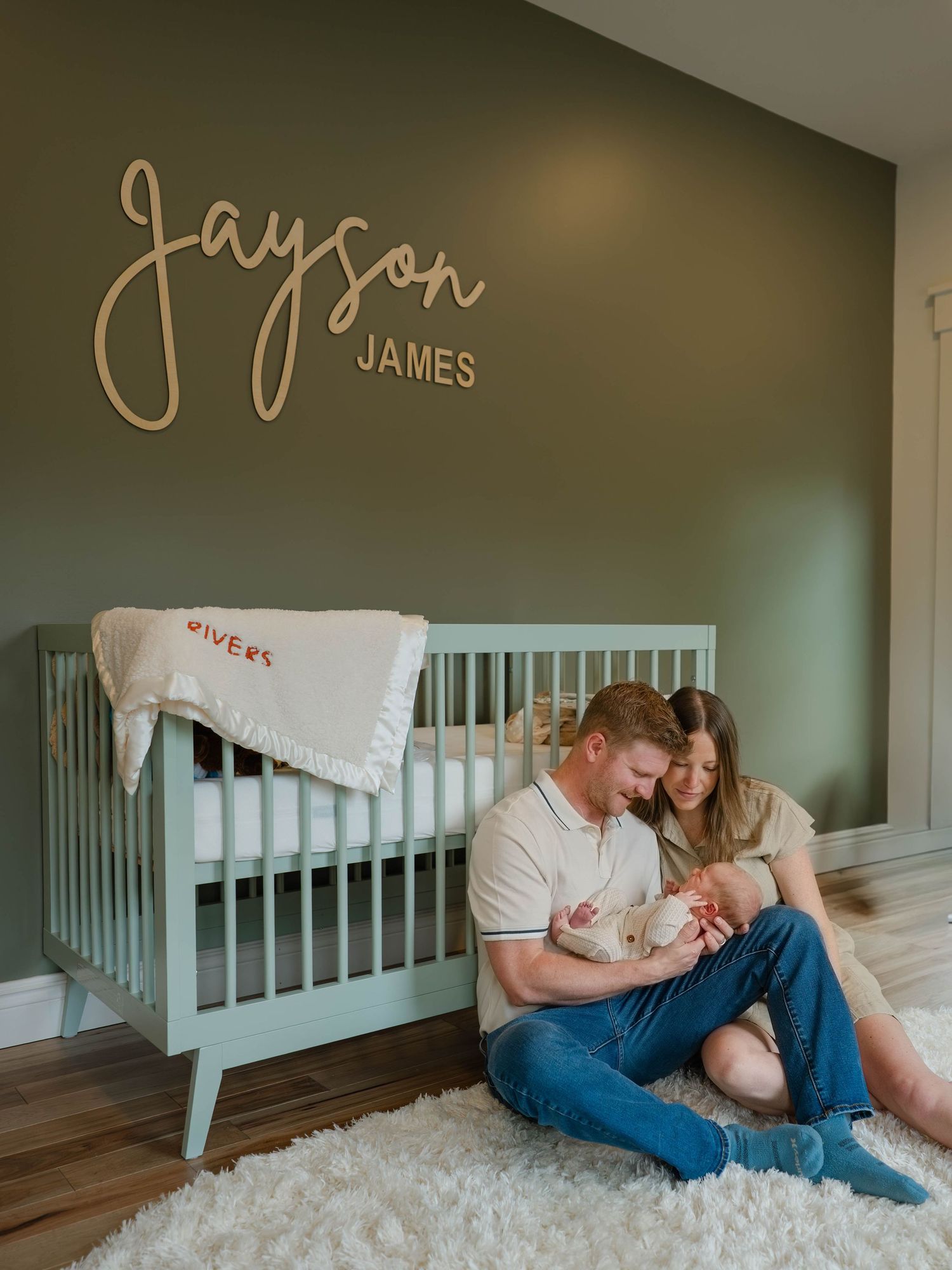 New parents sit on floor holding newborn in nursery with green walls and Jayson James wooden name sign above crib.