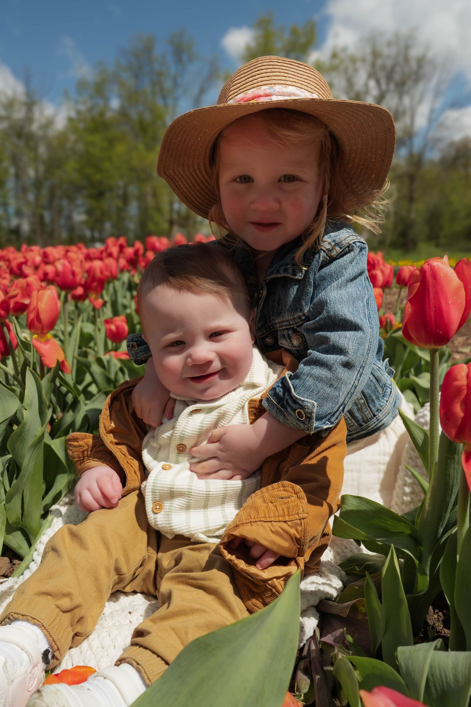 Sweet toddler in straw hat hugging smiling baby in a red tulip field.