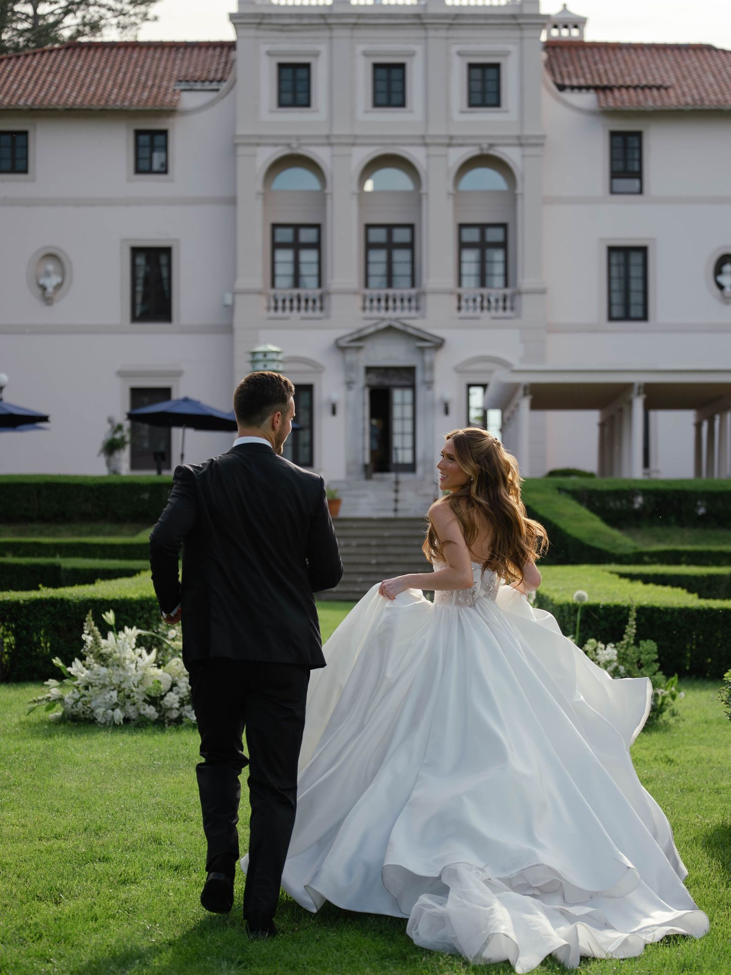 Bride and groom walking toward elegant white mansion wedding venue with manicured hedges and lush green lawn.