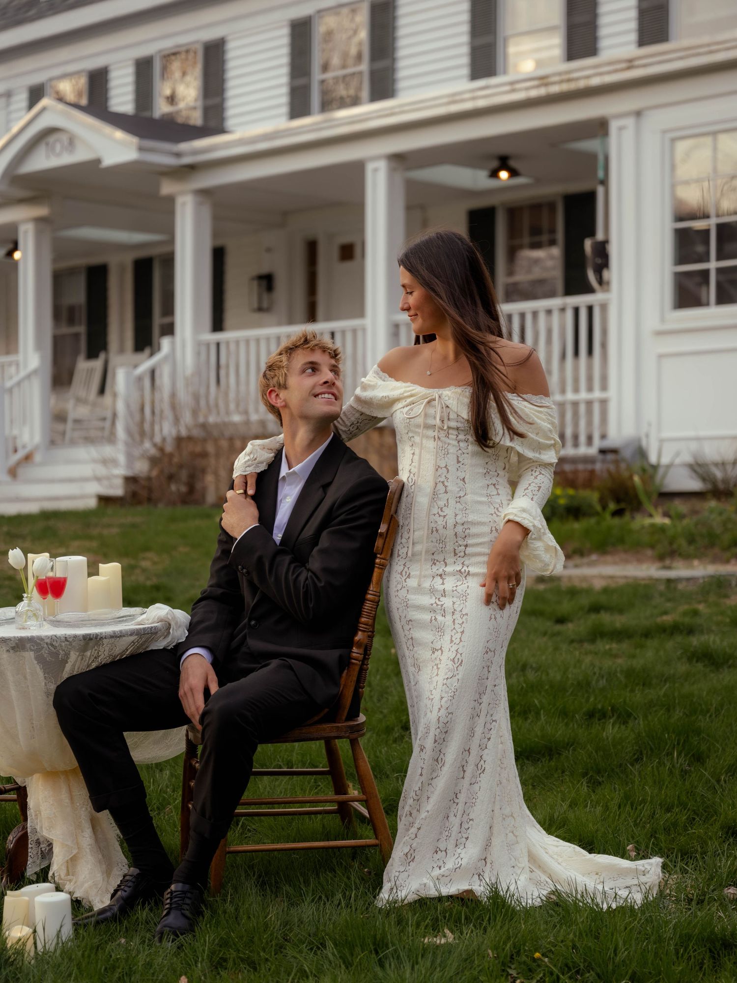 Bride in off-shoulder lace gown stands beside seated groom at candlelit table before white farmhouse.