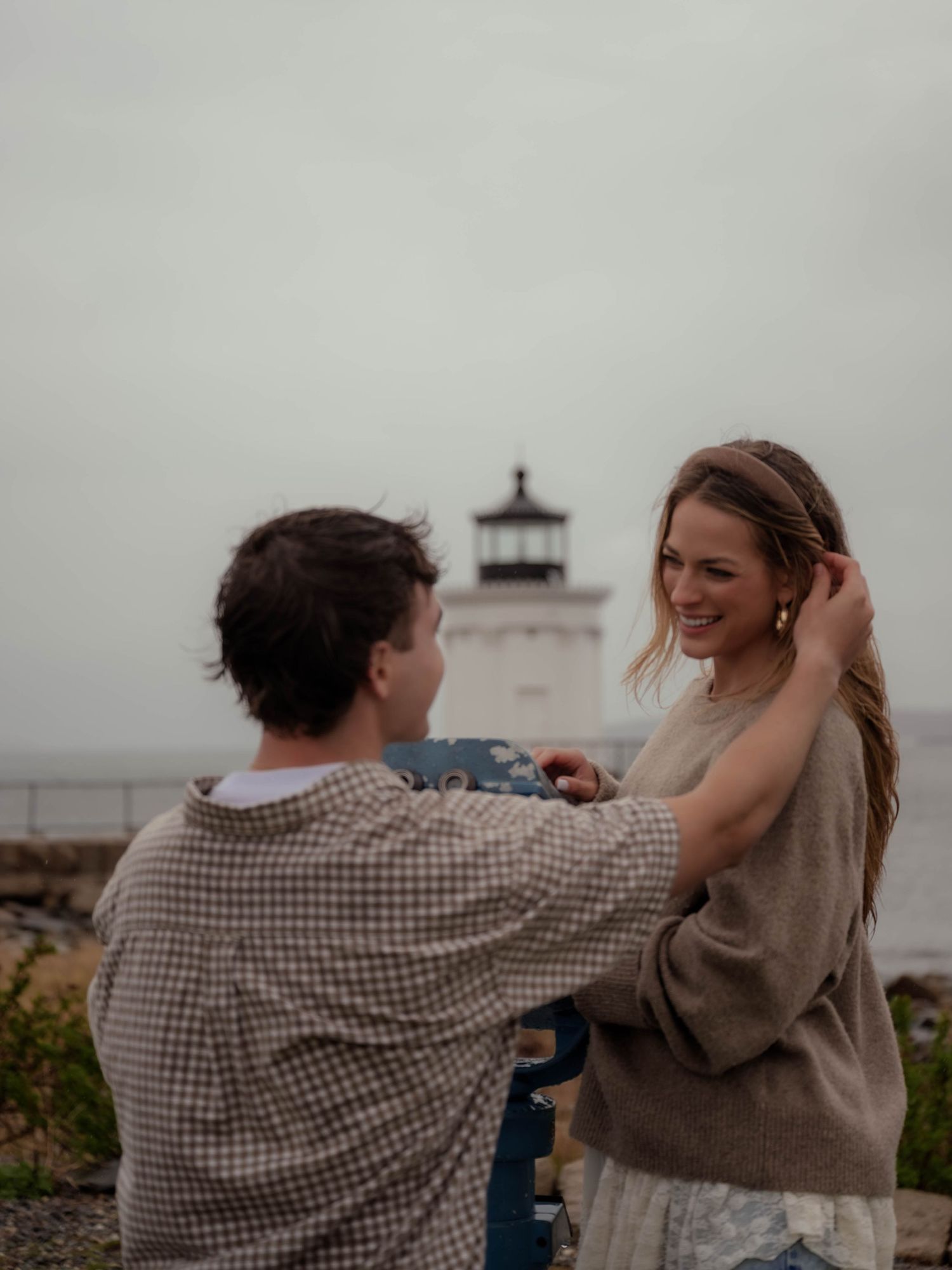 Man on one knee proposing to smiling woman near scenic coastal lighthouse.