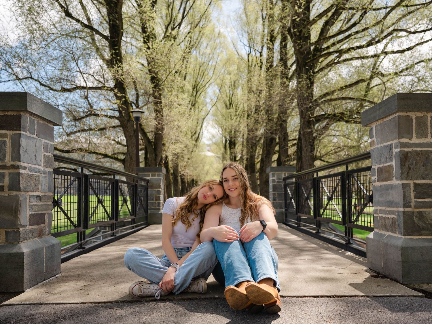 Two smiling young women sit together on a scenic tree-lined bridge pathway on a sunny day.