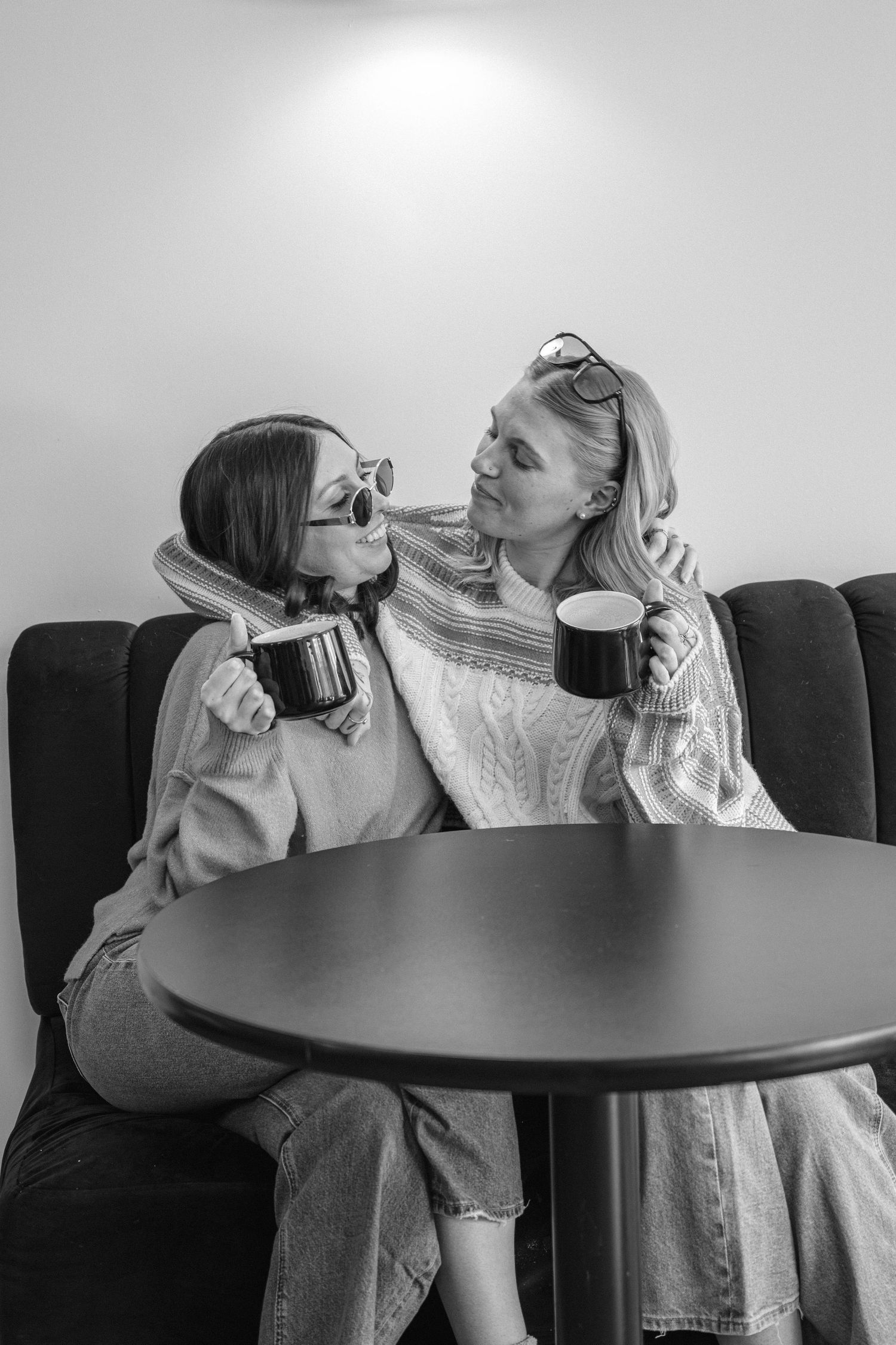 Black and white photo of two women sharing a close moment with coffee mugs at a cafe table.