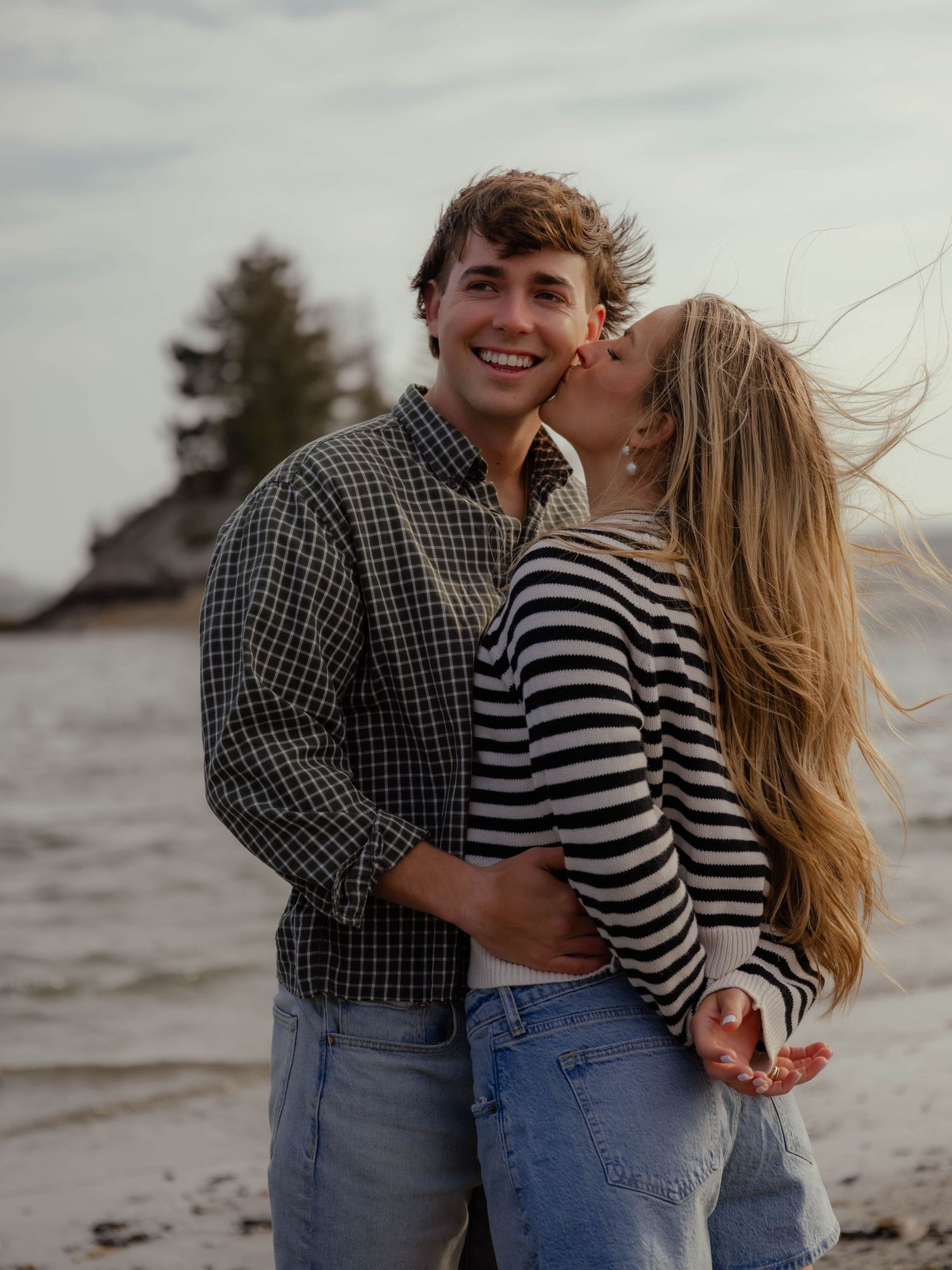 Happy couple on a beach, woman kissing mans cheek as he smiles at the camera.