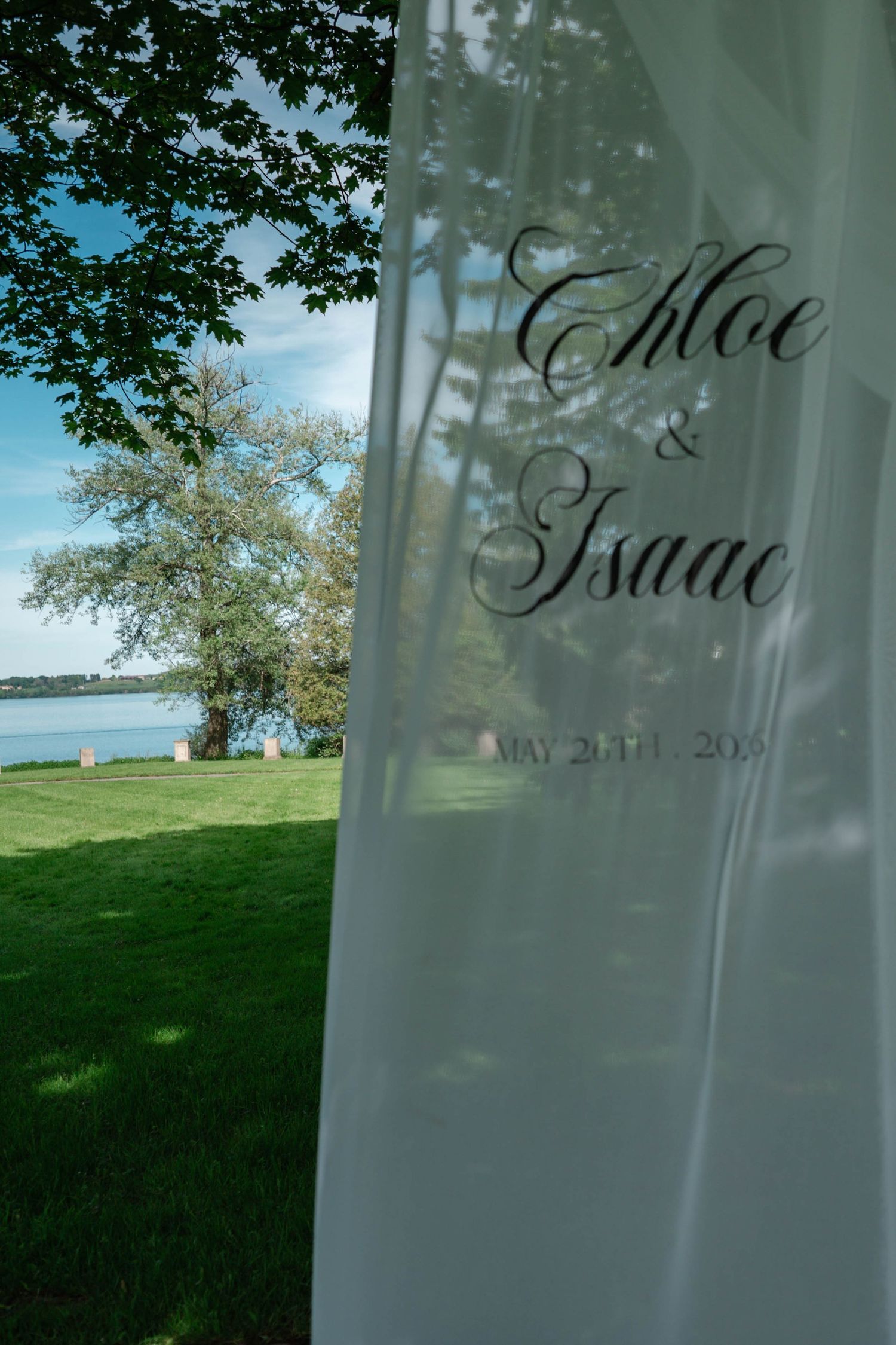 Wedding ceremony arch banner reading Chloe and Isaac May 20th 2025 with waterfront garden backdrop.