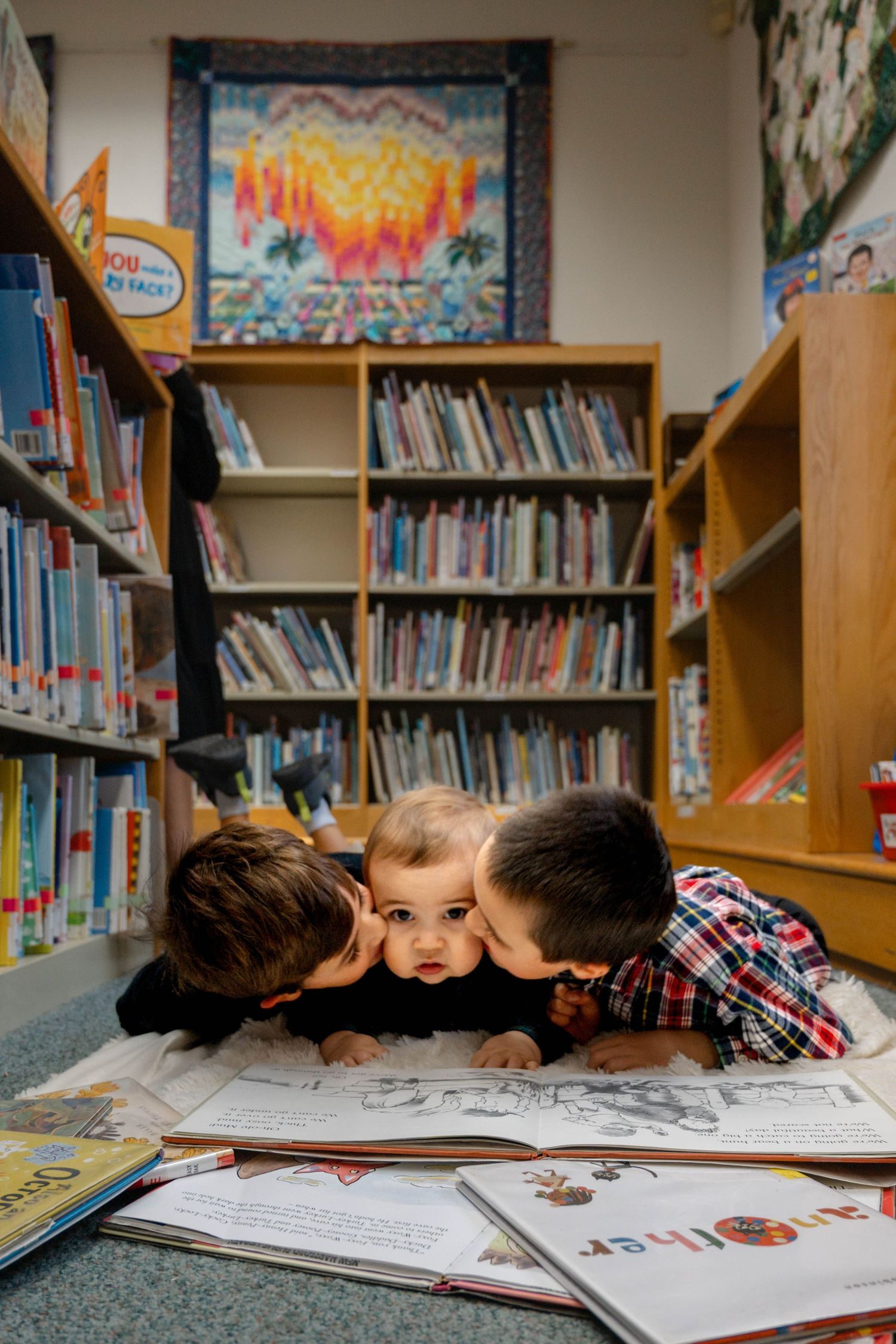 Two brothers kissing baby sibling on cheeks while lying on library floor surrounded by books.