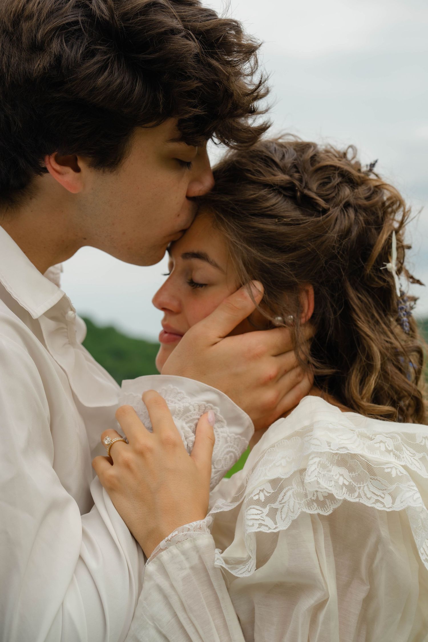 A groom tenderly kisses his bride on the forehead, both dressed in white, in a romantic outdoor setting.