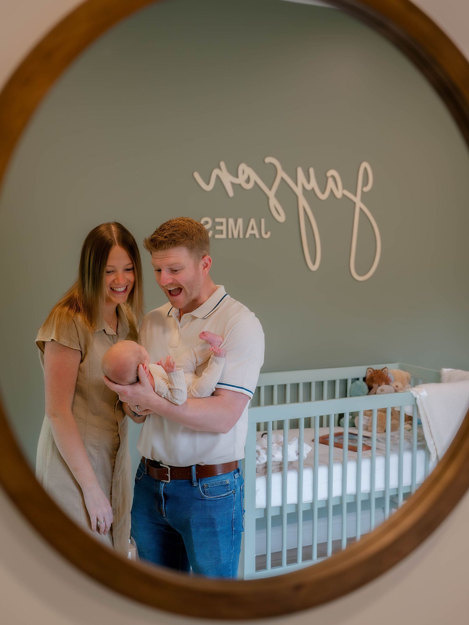 Parents smiling at newborn baby in nursery, reflected in round mirror with name sign on sage green wall.