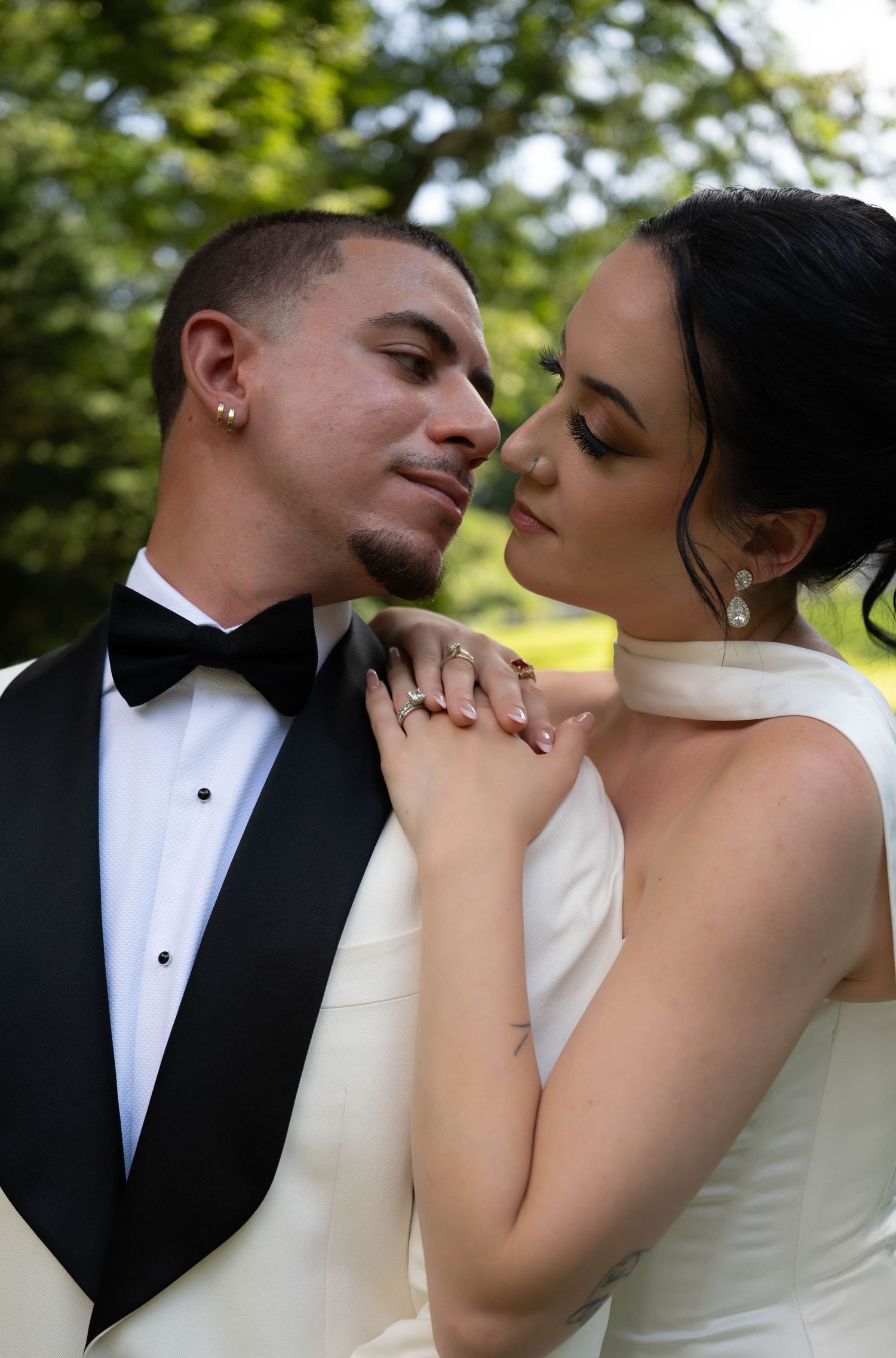 Bride and groom in elegant wedding attire share an intimate moment outdoors, with lush green trees in the background.