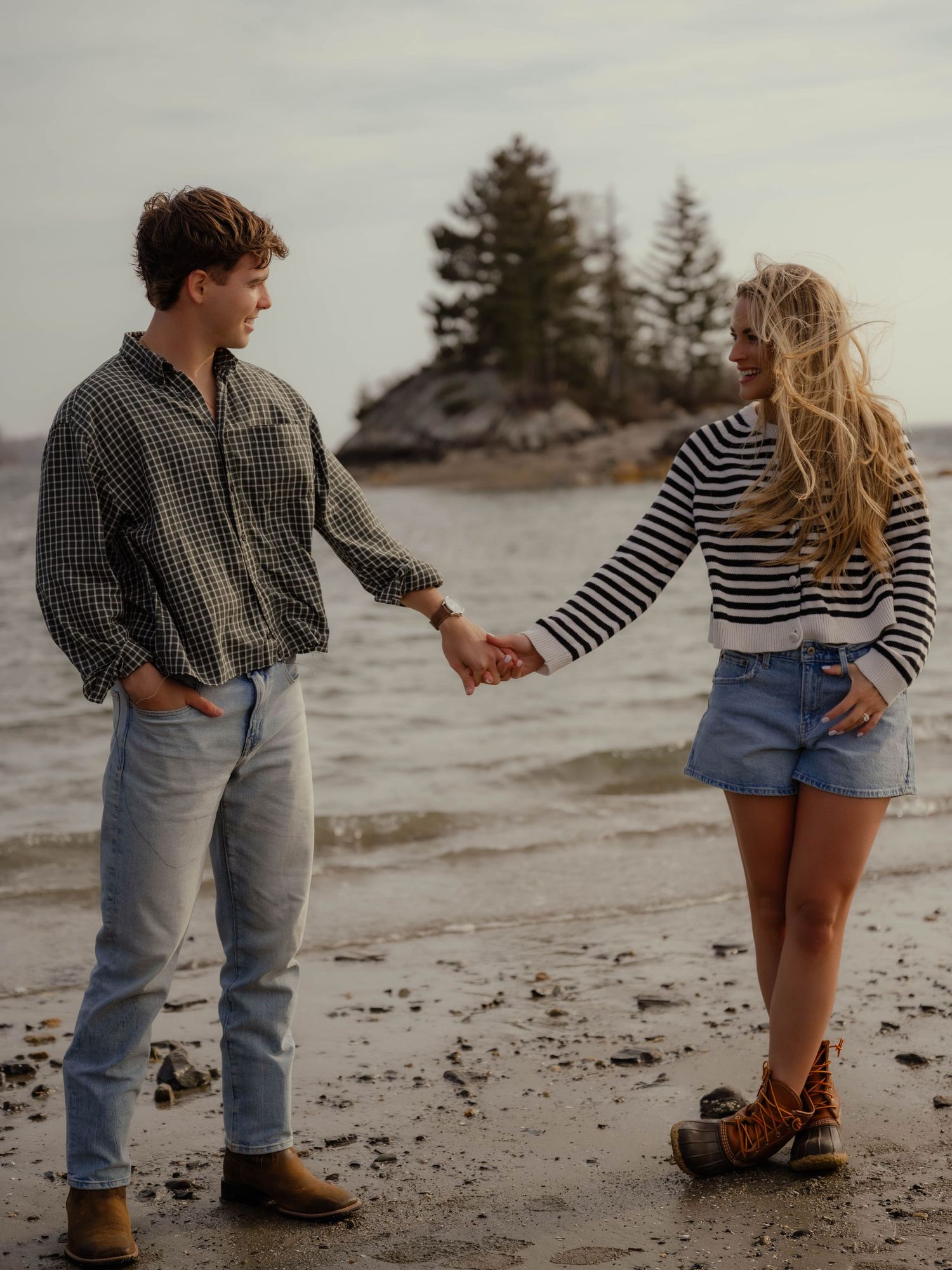 Young couple holding hands on a rocky beach with pine trees and water in the background.